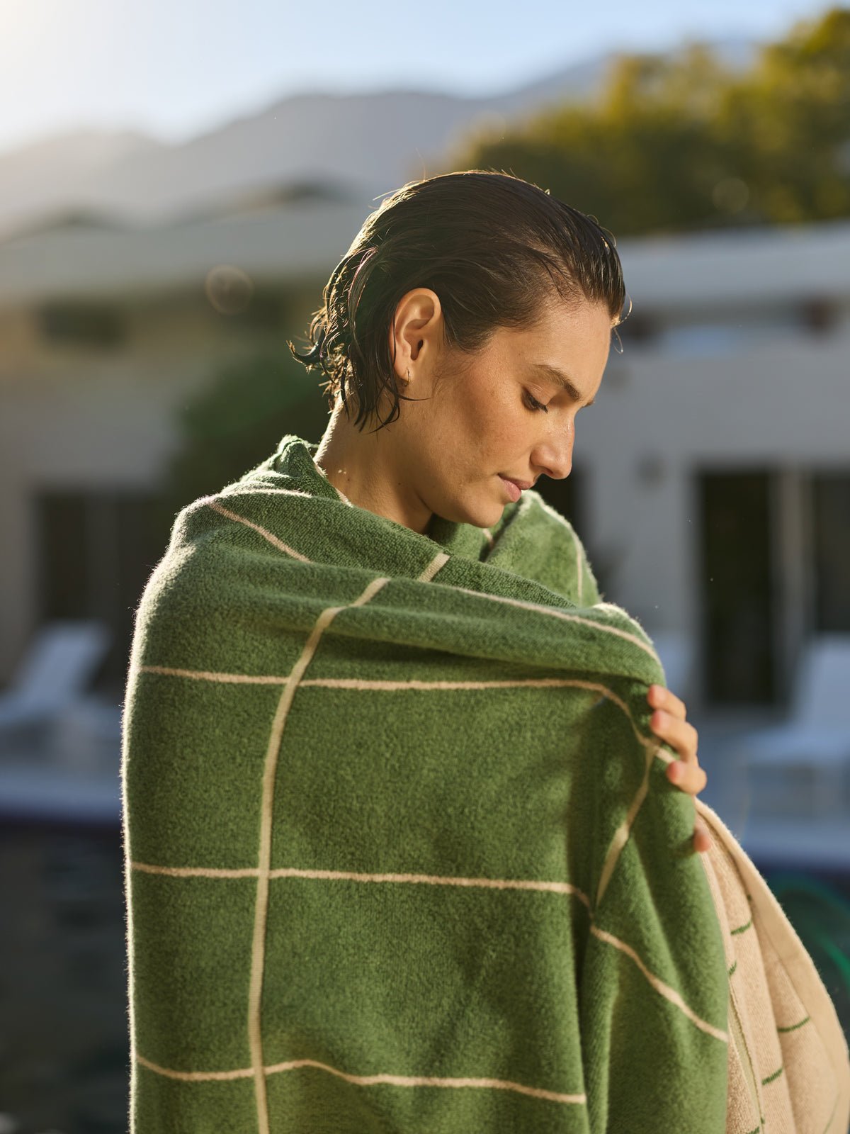 A person with wet hair stands wrapped in a Cozy Earth Windowpane Resort Towel, looking downwards. The background shows a blurred house and trees with sunlight filtering through. 