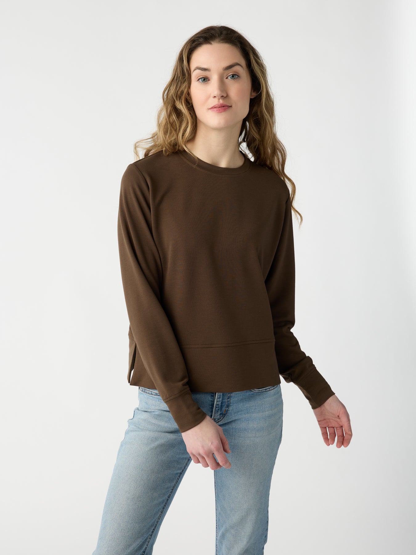 Someone with long, wavy hair wears a Cozy Earth Women's StudioLite Crewneck and light blue jeans, standing against a plain white background while looking directly at the camera with a neutral expression. |Color:Sable