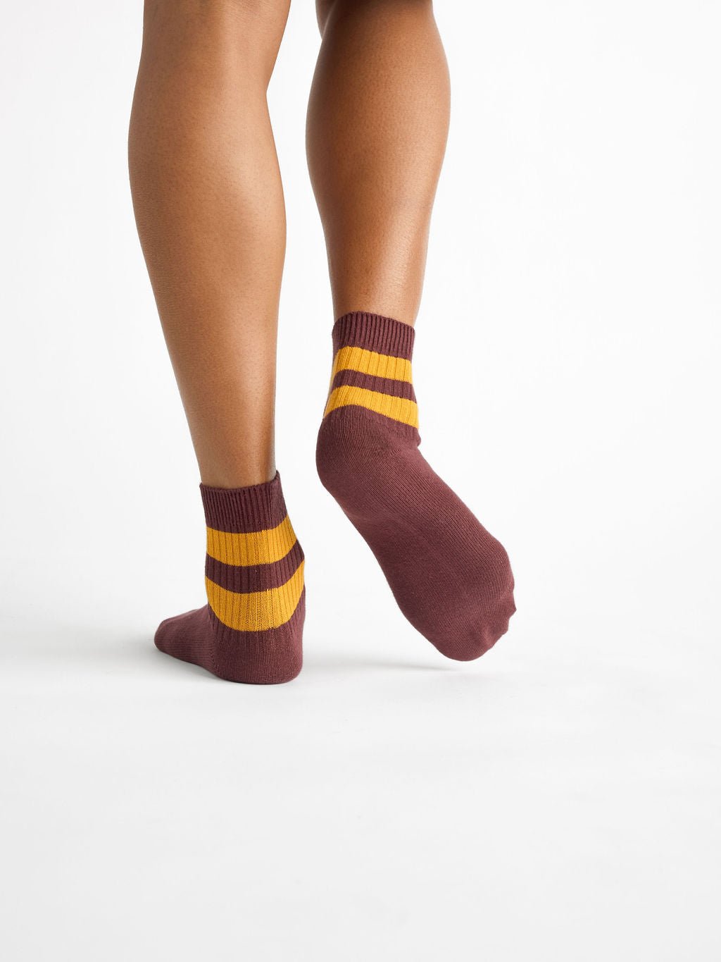 A person stands on one foot showing the back of Cozy Earth's Women's Sadie Sock in maroon with two wide yellow stripes near the top, set against a plain white background. 