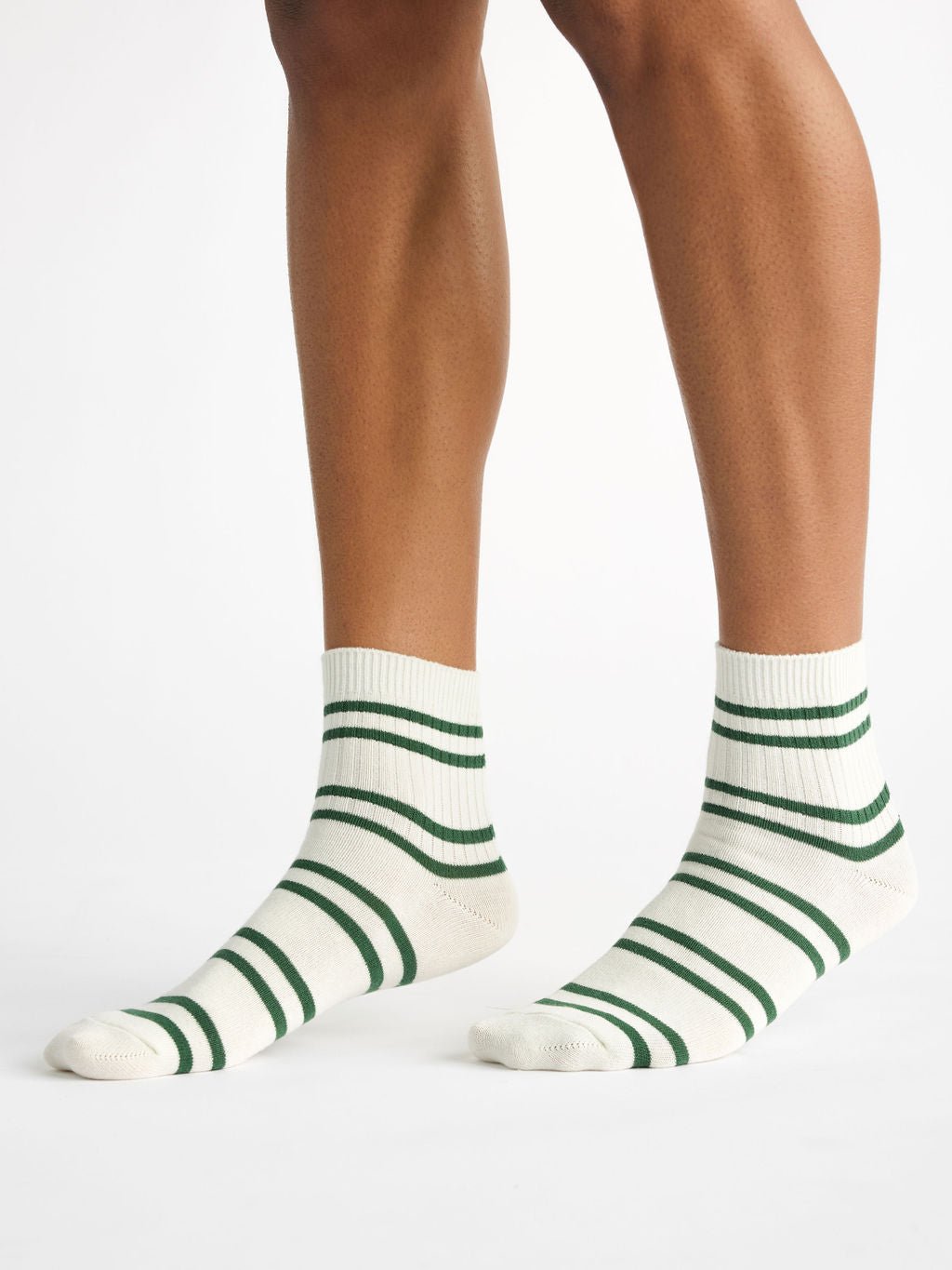 A person wears the Women's Sadie Sock by Cozy Earth, featuring cream-colored fabric with green horizontal stripes, on a white background. The view shows legs from below the knee to the feet. 