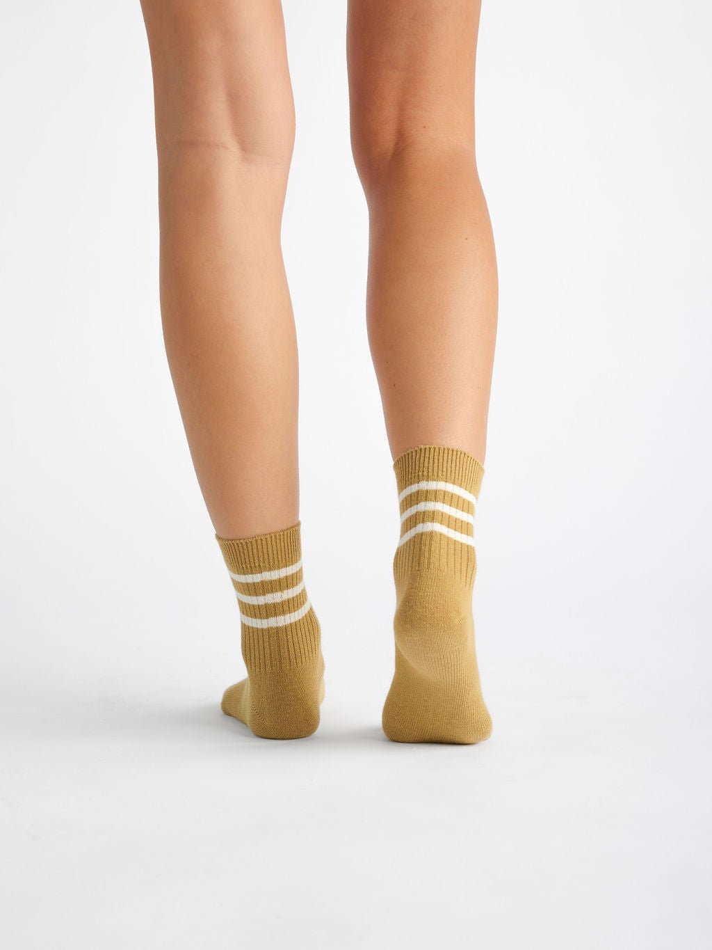 Back view of someone wearing Cozy Earth's Women's Sadie Socks in mustard yellow with two white stripes near the top, mid-calf height, standing on a plain white background. 