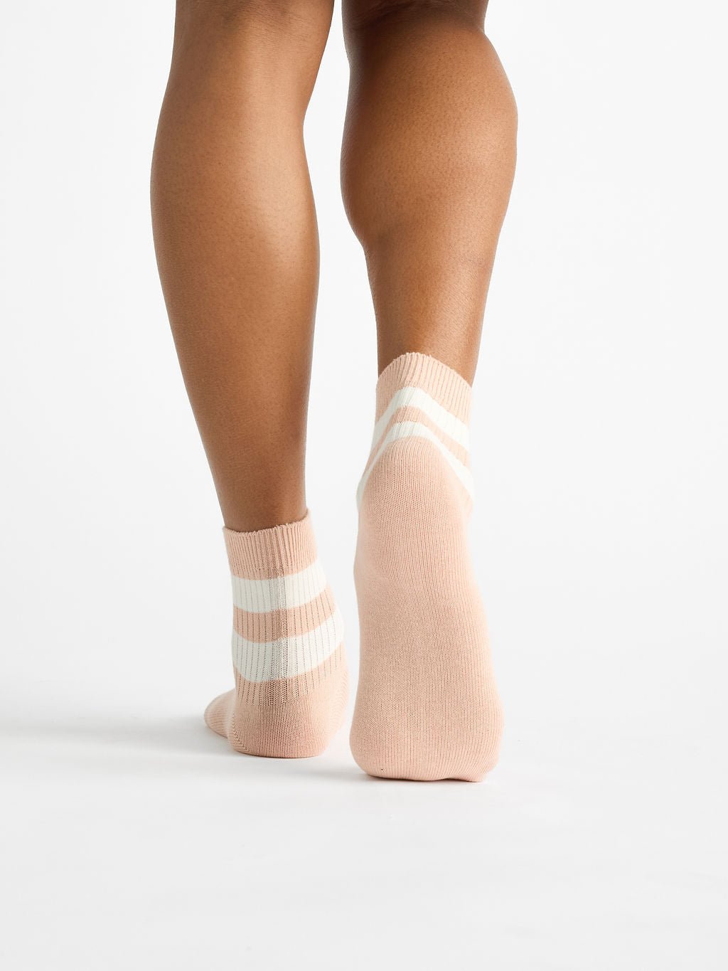 A person is seen from the rear wearing Cozy Earth's Women's Sadie Socks, which are ankle-length and beige with white stripes and ribbed texture, against a plain white background. 