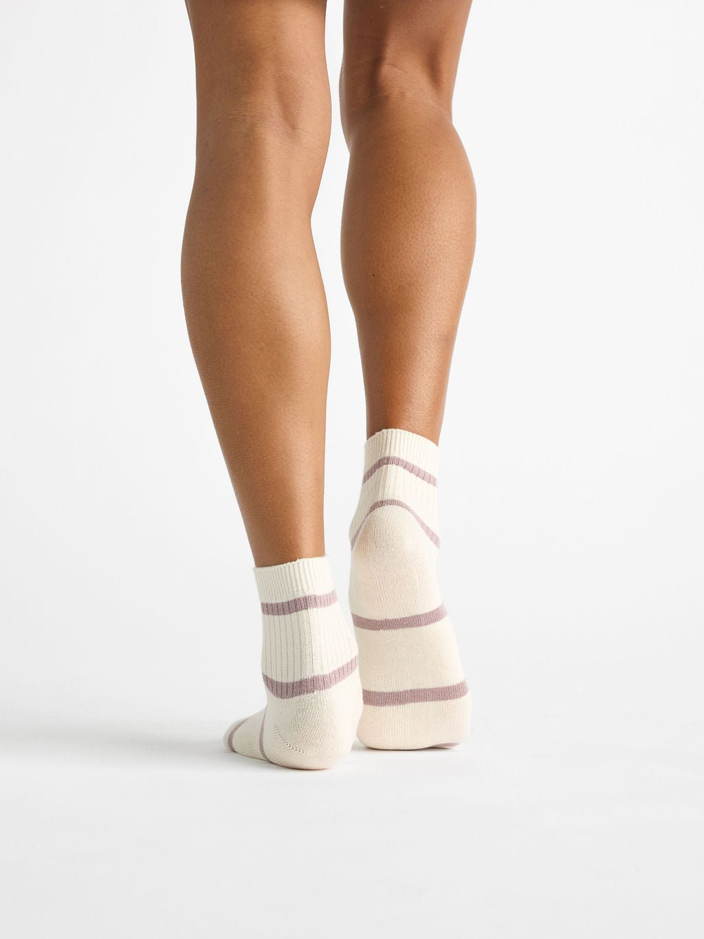 A person stands with their back to the viewer, wearing Women's Sadie Sock by Cozy Earth, featuring cream-colored fabric with light purple stripes. The white floor and background create a minimalistic setting. 