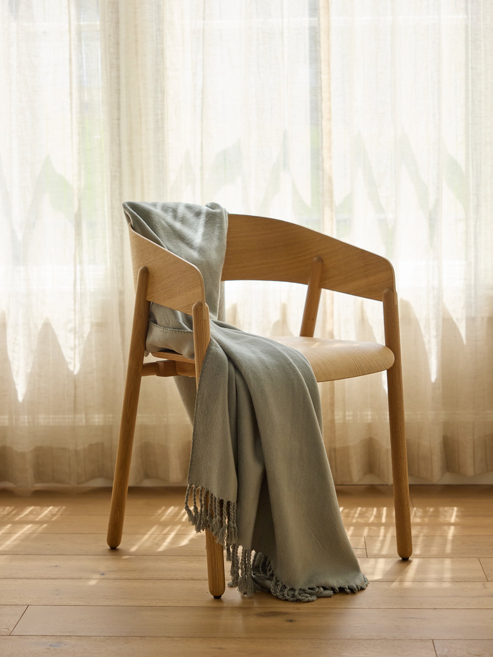 A wooden chair with a curved backrest and armrests is placed in front of sheer, white curtains. Draped over the chair's arm is the Cozy Earth Bamboo Tassel Throw, a light gray blanket with fringed edges, cascading down to the wooden floor. Sunlight filters gently through the curtains. 