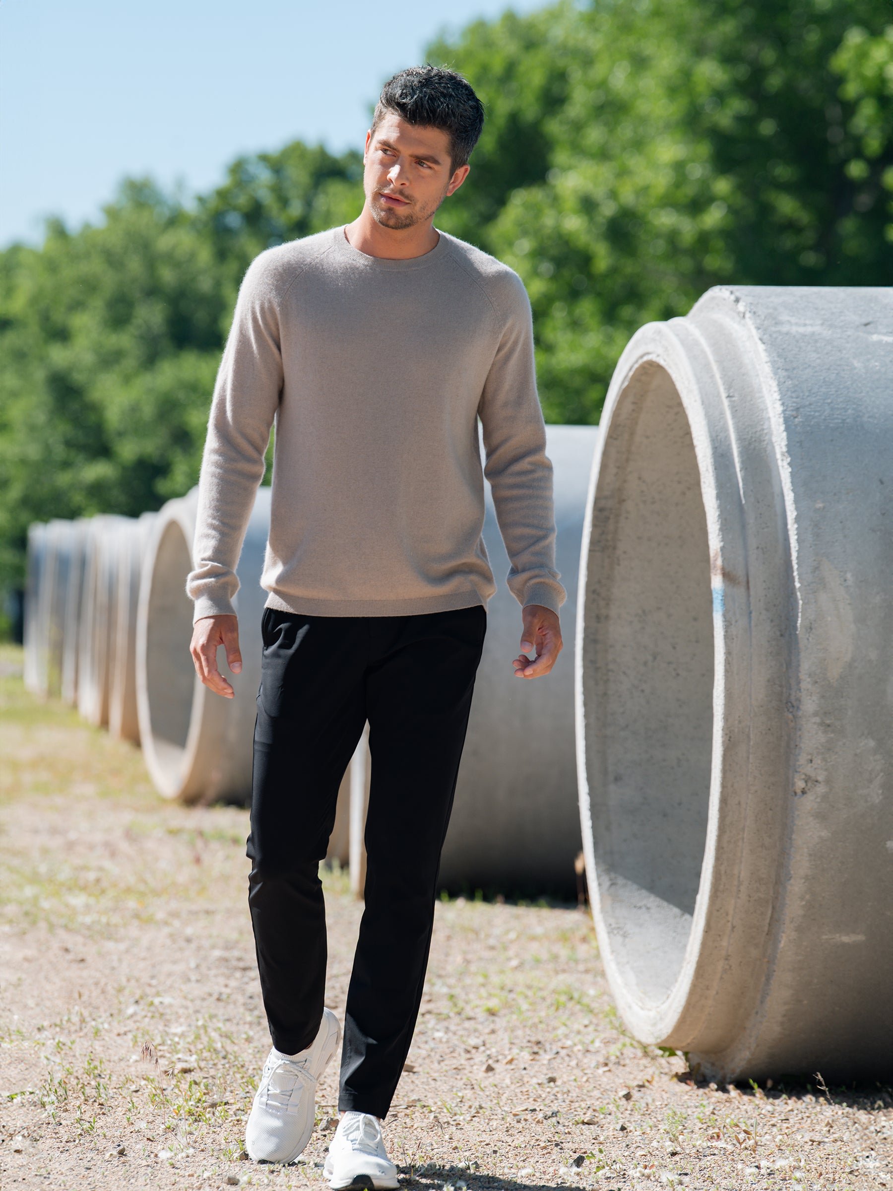 A man wearing a Cozy Earth Men's Crewneck Sweater in beige and black pants walks outdoors near large concrete pipes. He has dark hair and facial hair. Trees and a blue sky are visible in the background. As he strolls along a gravel path, he glances slightly to his left. 