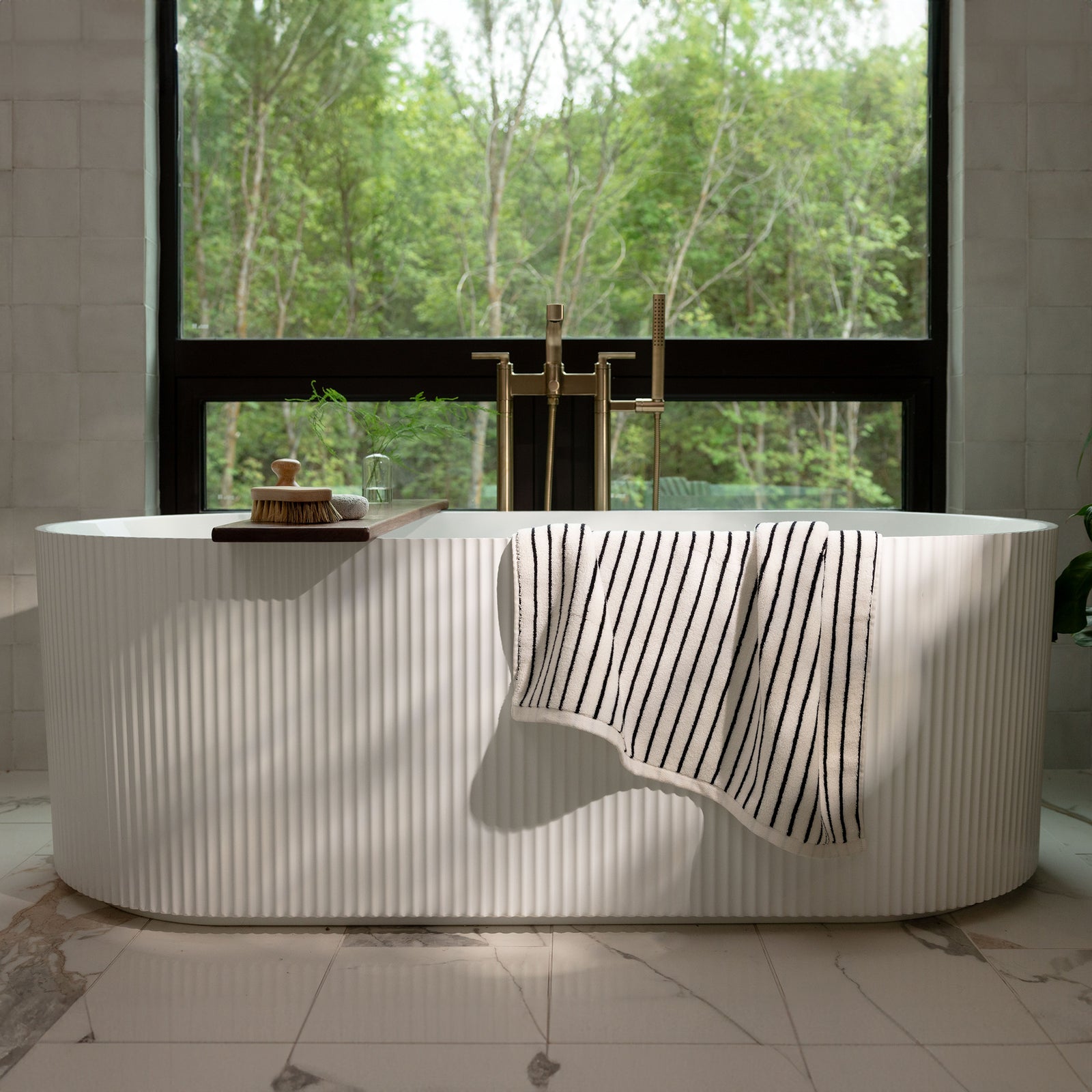 A serene bathroom features a white freestanding bathtub with a Luxe Bath Towel from Cozy Earth draped over the side. Behind the tub, a large window showcases a lush green forest, allowing natural light to flood the space. The floor is made of white marble with subtle grey veins. 