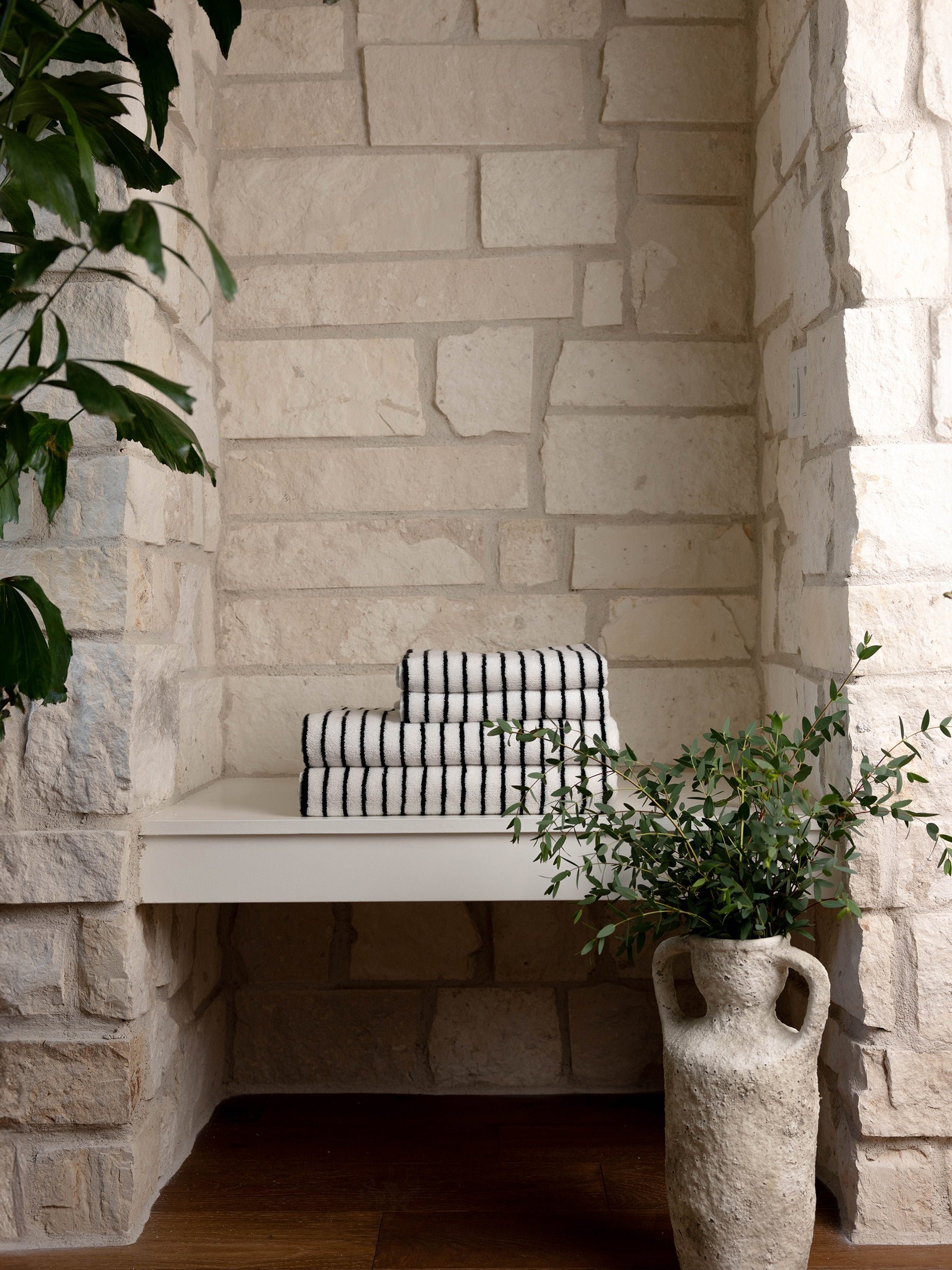 A neatly stacked pile of folded Luxe Hand Towels by Cozy Earth, featuring black and white stripes, sits on a white shelf against a textured stone wall. A rustic vase filled with green leafy branches is placed on the floor next to the shelf. A large plant with dark green leaves is partially visible on the left. 