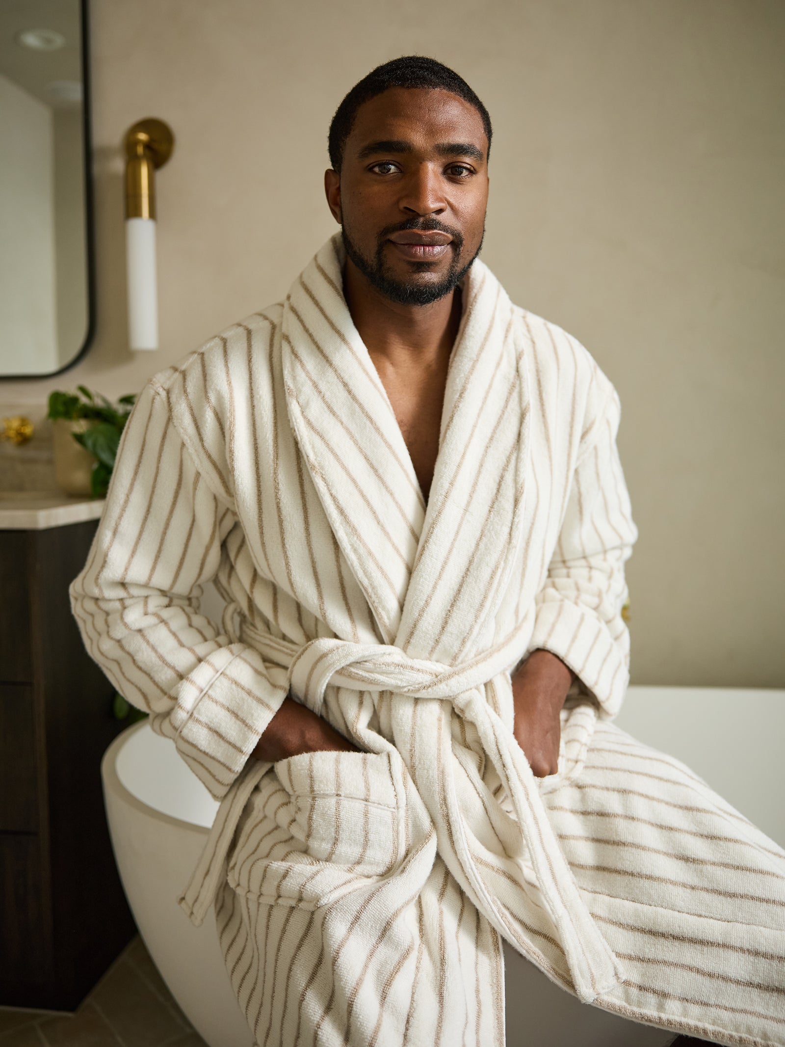 A person with short hair and a beard sits on the edge of a bathtub wearing the Luxe Bath Robe from Cozy Earth, which is cream-colored with stripes. The background features a wall sconce along with partial views of a mirror and cabinet. 