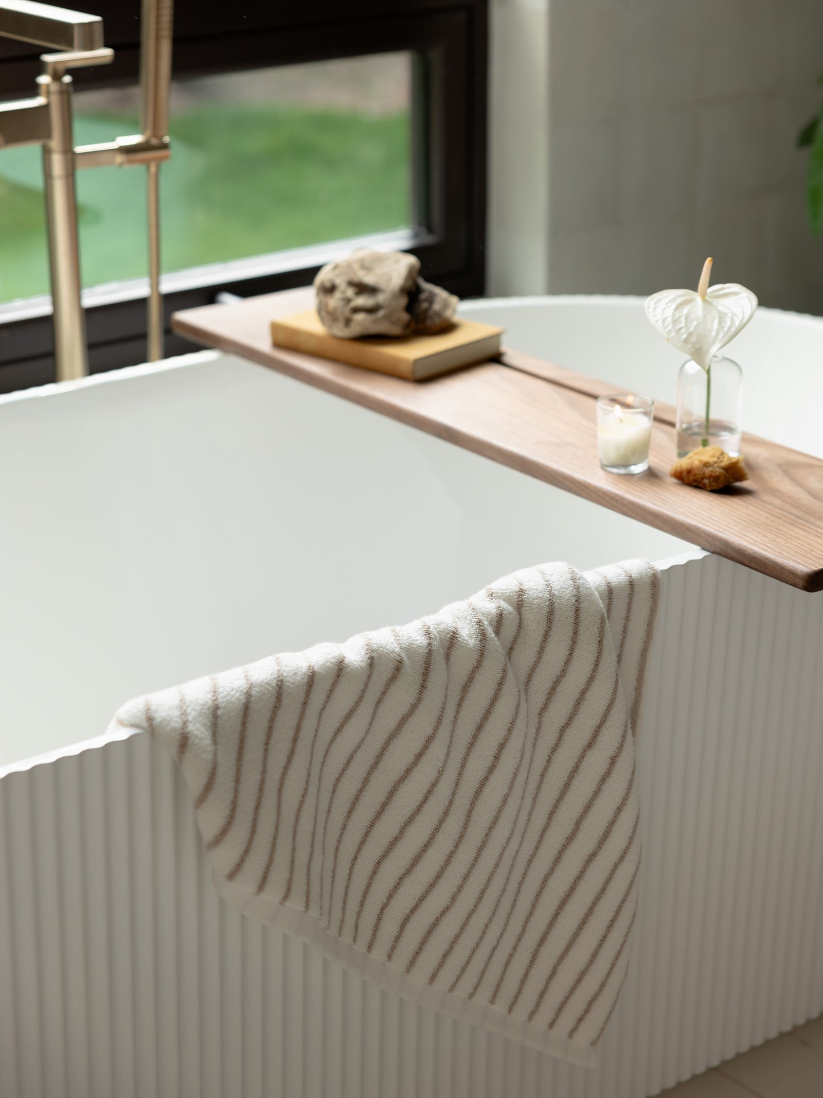 A modern white bathtub with a textured exterior stands near a window. A wooden tray spans the tub, holding two glass vases with plants, a candle, and a decorative stone. Draped over the tub's edge is a Luxe Bath Towel featuring thin, wavy brown stripes from Cozy Earth. 