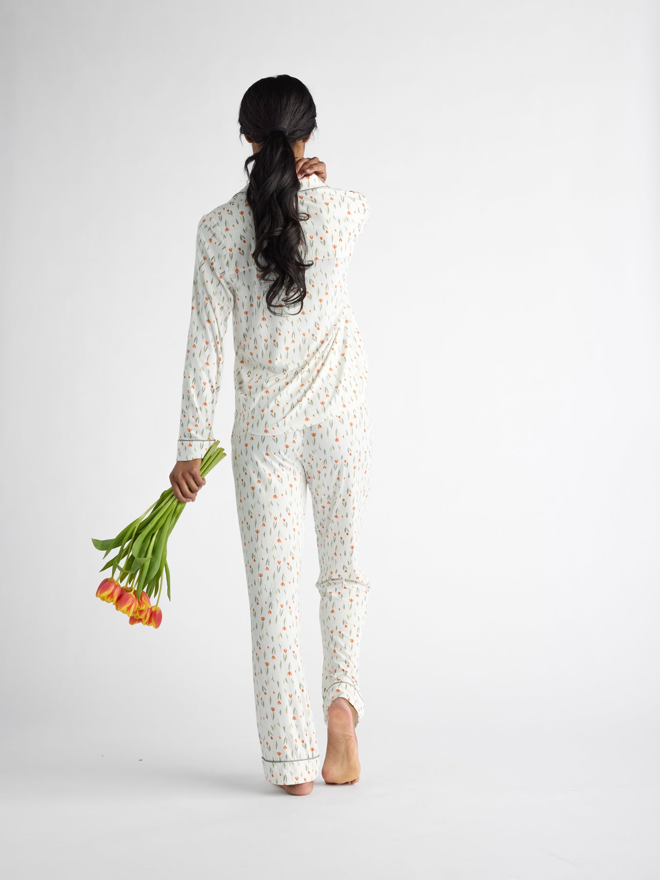 A long-haired person stands barefoot, turned away, wearing Cozy Earth Women's Bamboo Stretch-Knit Pajama Pants. They hold a bouquet of orange tulips in their right hand against a plain white backdrop. 