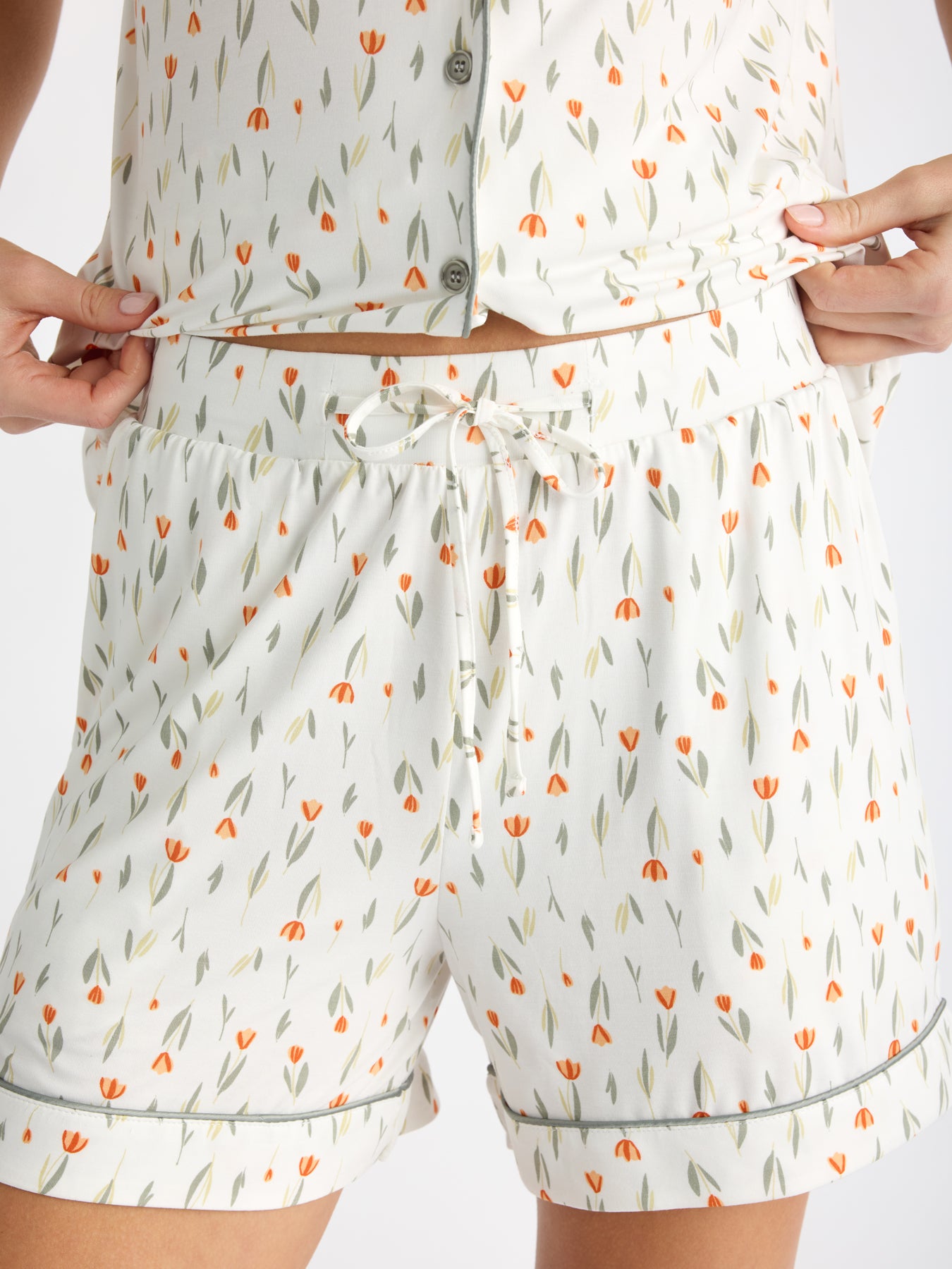 A person wearing HIDE's Women's Bamboo Stretch-Knit Pajama Short in a white floral pattern with small orange tulips adjusts their button-up top, revealing the waistband of the shorts. 