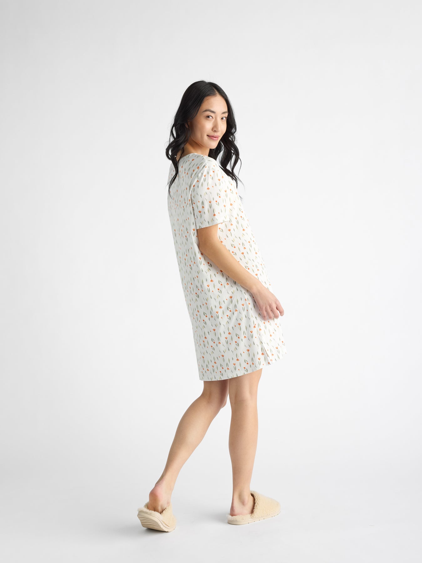 A person with long hair smiles slightly, modeling the Cozy Earth Women's Bamboo Stretch-Knit Sleep Dress, featuring a colorful pattern. They wear cozy slippers and stand turned slightly to the side against a plain white backdrop. 