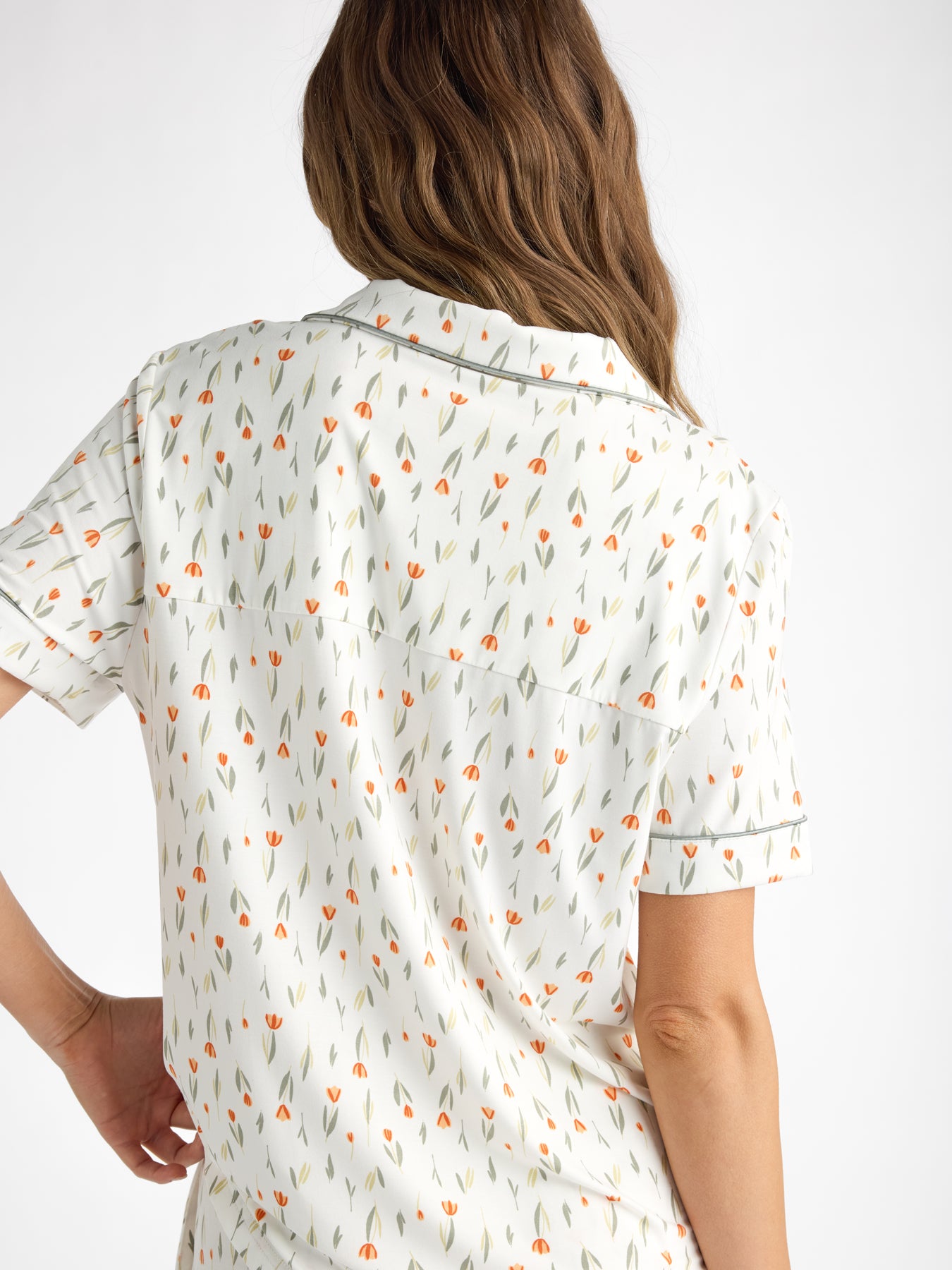 A person with long wavy brown hair wears a HIDE Women's Bamboo Stretch-Knit Short Sleeve Pajama Top, featuring a small orange and green floral pattern on a white background. 