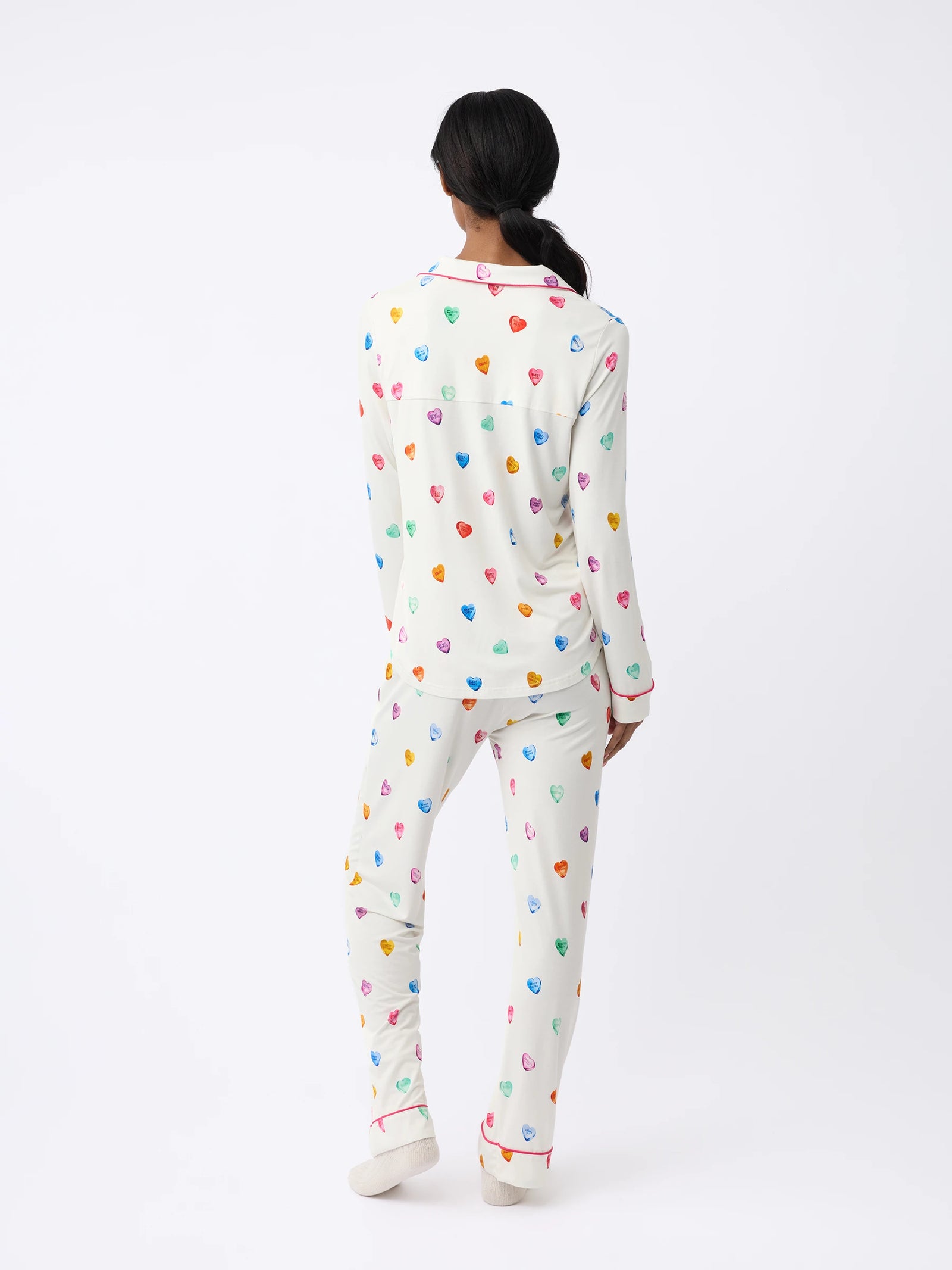 A person stands against a plain white background wearing the Women's Long Sleeve Bamboo Pajama Top by Cozy Earth, with colorful heart patterns and a collar. The bottoms reach the ankles. 