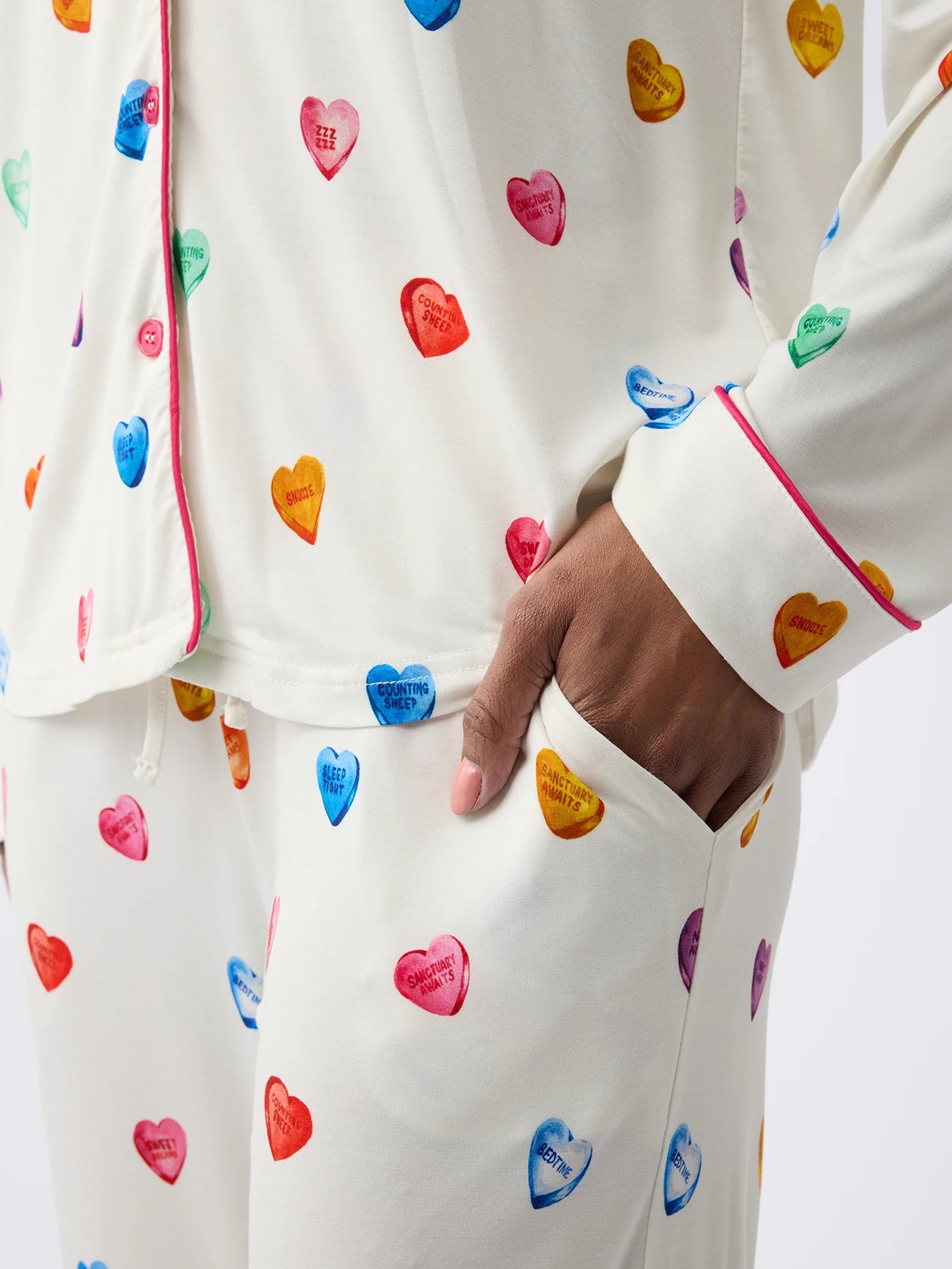 Someone wears Cozy Earth Women's Stretch-Knit Bamboo Pajama Pant in cream with colorful heart patterns and phrases like "BE MINE" and "HEY GIRL," adorned with red piping on the top. One hand is casually placed in the pant pocket. 