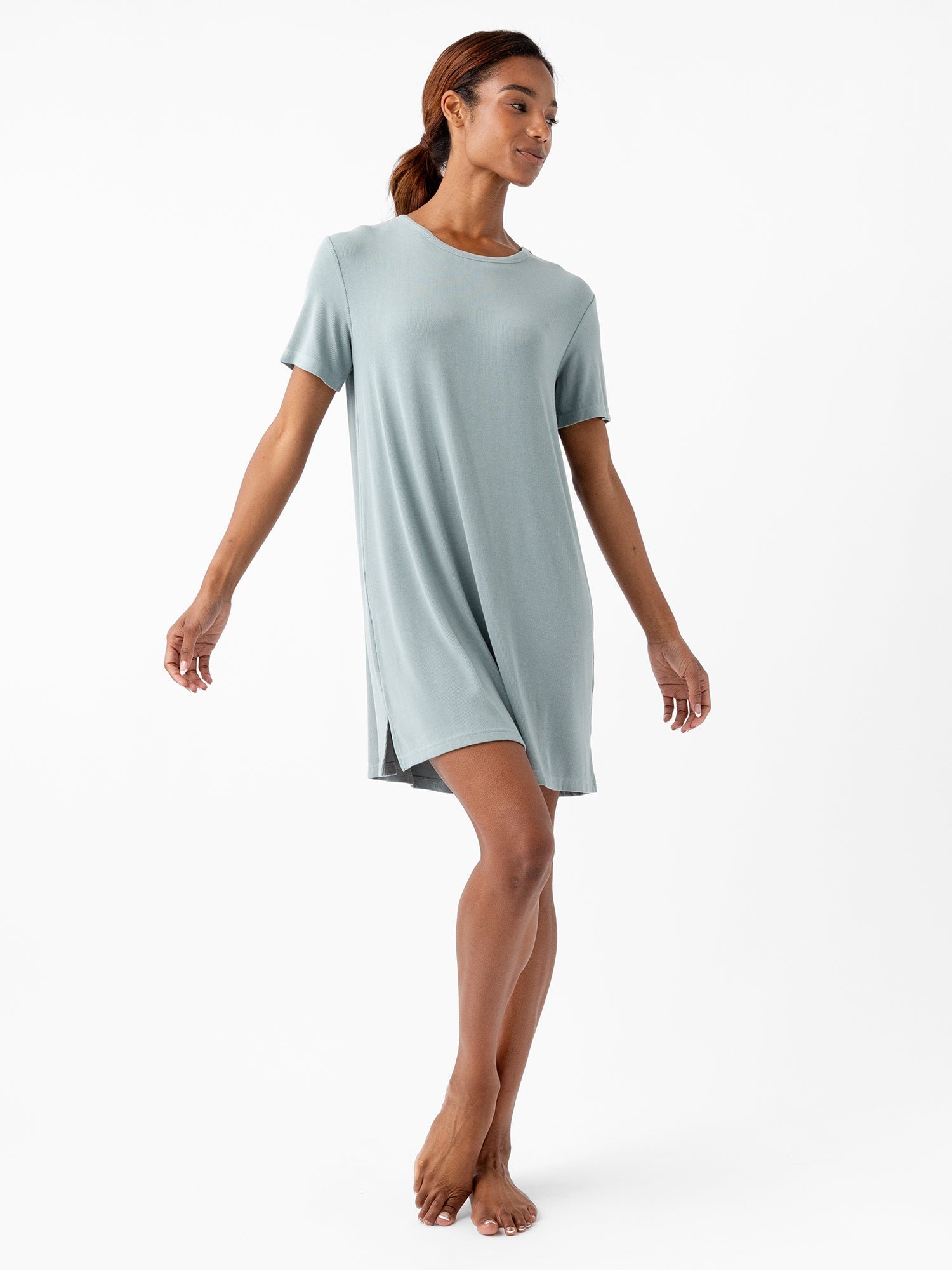A woman stands barefoot on a white background, wearing the loose-fitting, short-sleeved Women’s Bamboo Rib-Knit Boyfriend Sleep Dress by Cozy Earth. Her head is slightly turned to the side, and her arms are extended, giving a sense of balance and grace. The teal sleep dress features a small slit on one side. 