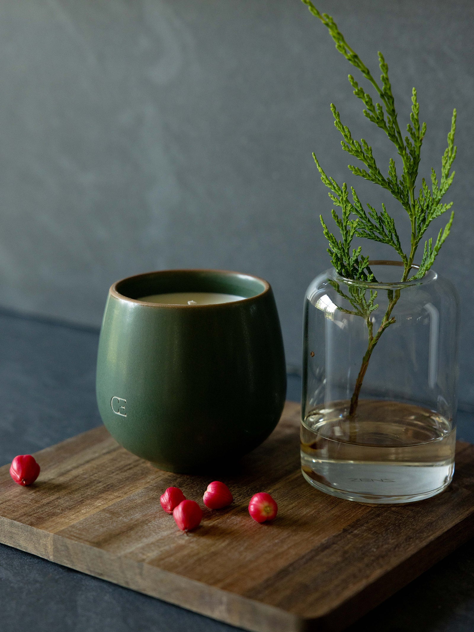 Cozy Earth Solstice Candle sits on a wooden board next to a vase with a sprig of evergreen