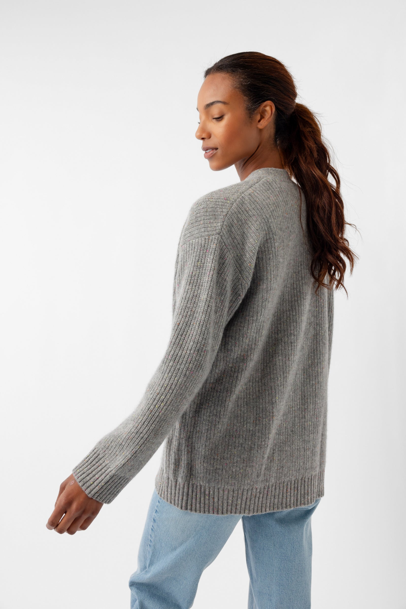 A person with long hair, dressed in an Oversized Sunday Cashmere Cardigan by Cozy Earth and light blue jeans, is standing slightly turned to the side, looking with a neutral expression against a plain white backdrop. 