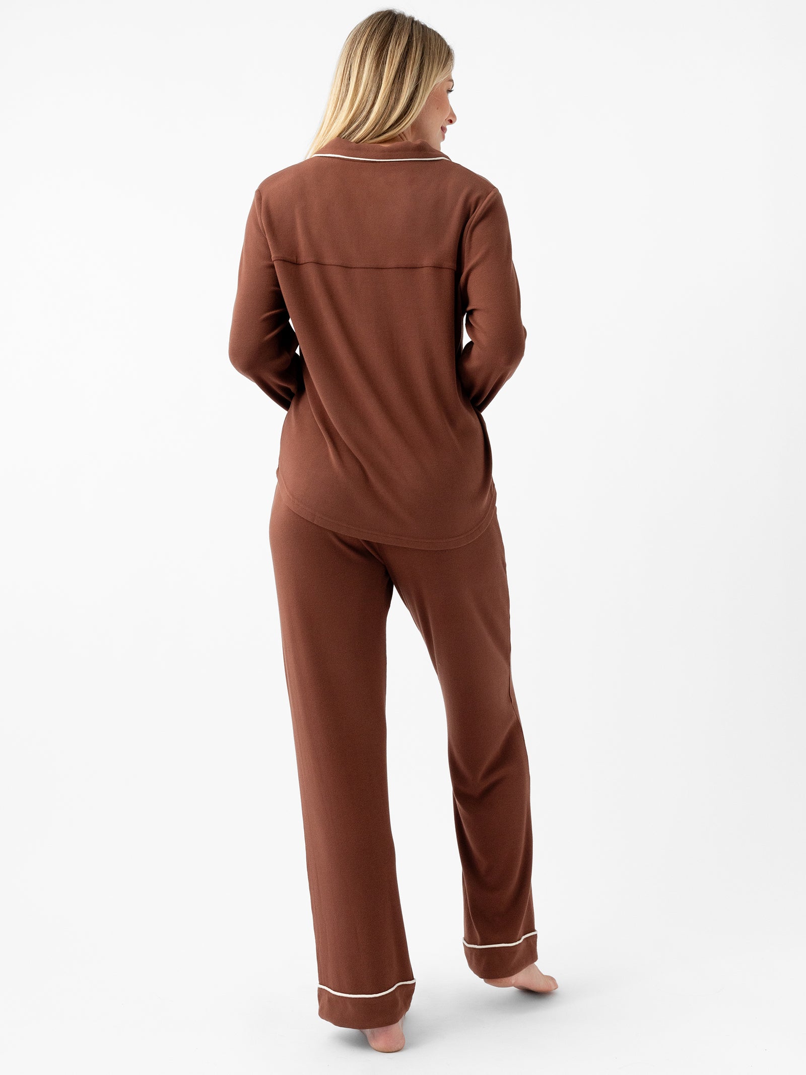 A person with long blonde hair is standing barefoot facing away from the camera, wearing a brown long-sleeve pajama top and matching Cozy Earth's Women's Bamboo Rib Knit Classic Pajama Pant with white trim on the cuffs. The background is plain white. 