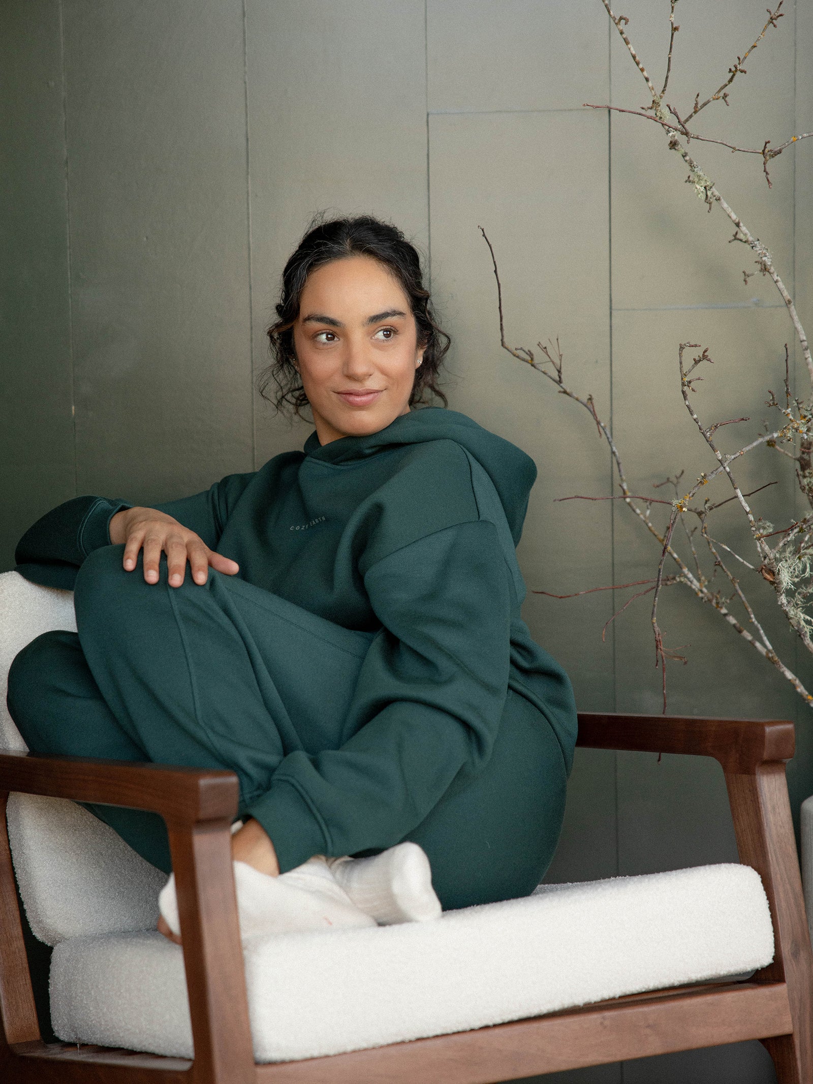 A person with dark hair sits comfortably on a white cushioned chair, wearing the Cozy Earth's Women's CityScape Hoodie paired with sweatpants. They have a relaxed expression as they gaze off-camera. In the background, a bare branch decorates the scene against a greenish wall. 