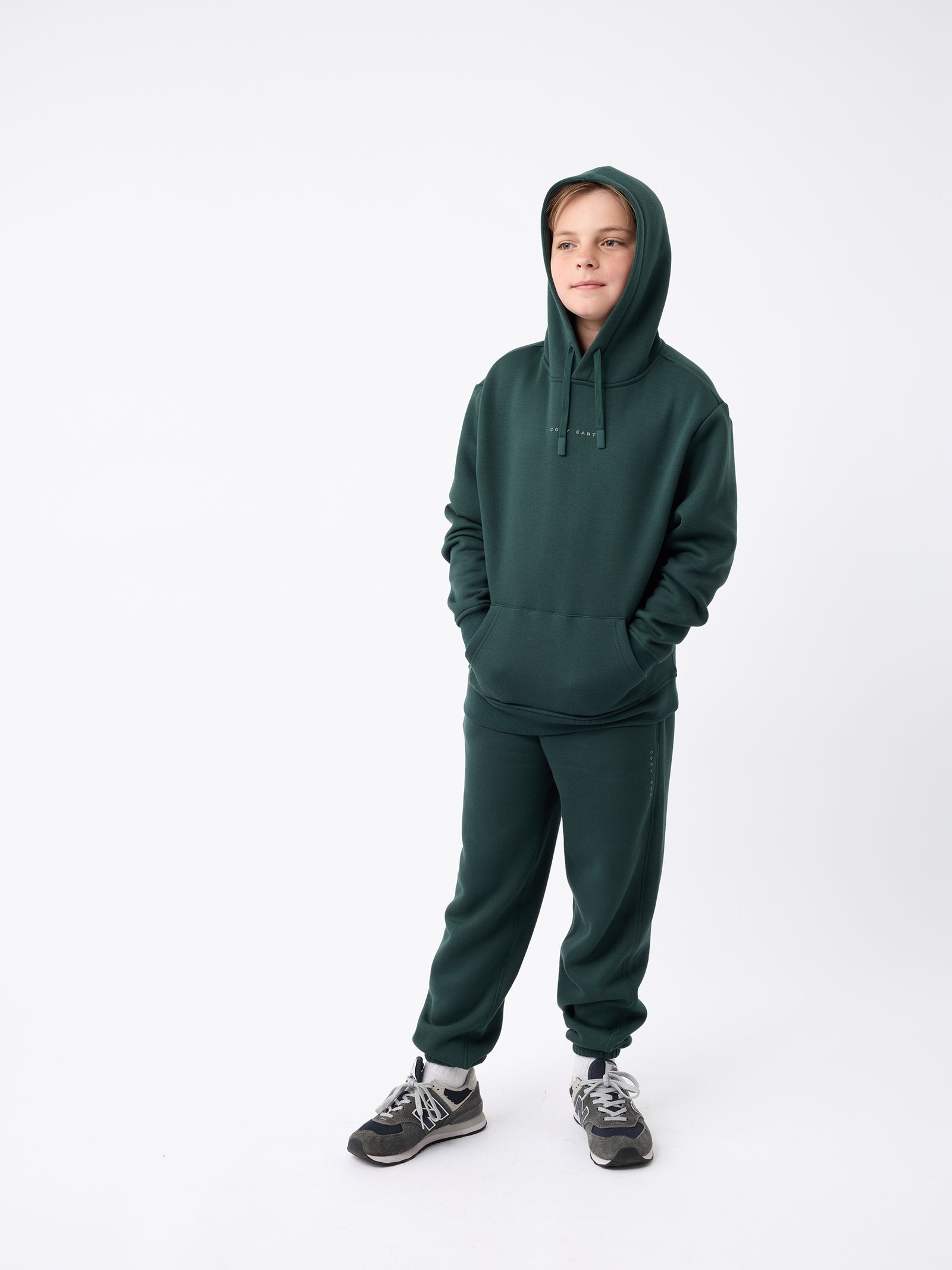 A young person is standing against a white background, dressed in a dark green Kid's CityScape Hoodie by Cozy Earth along with matching sweatpants. Their hands are in their pockets, and they're sporting gray sneakers. 