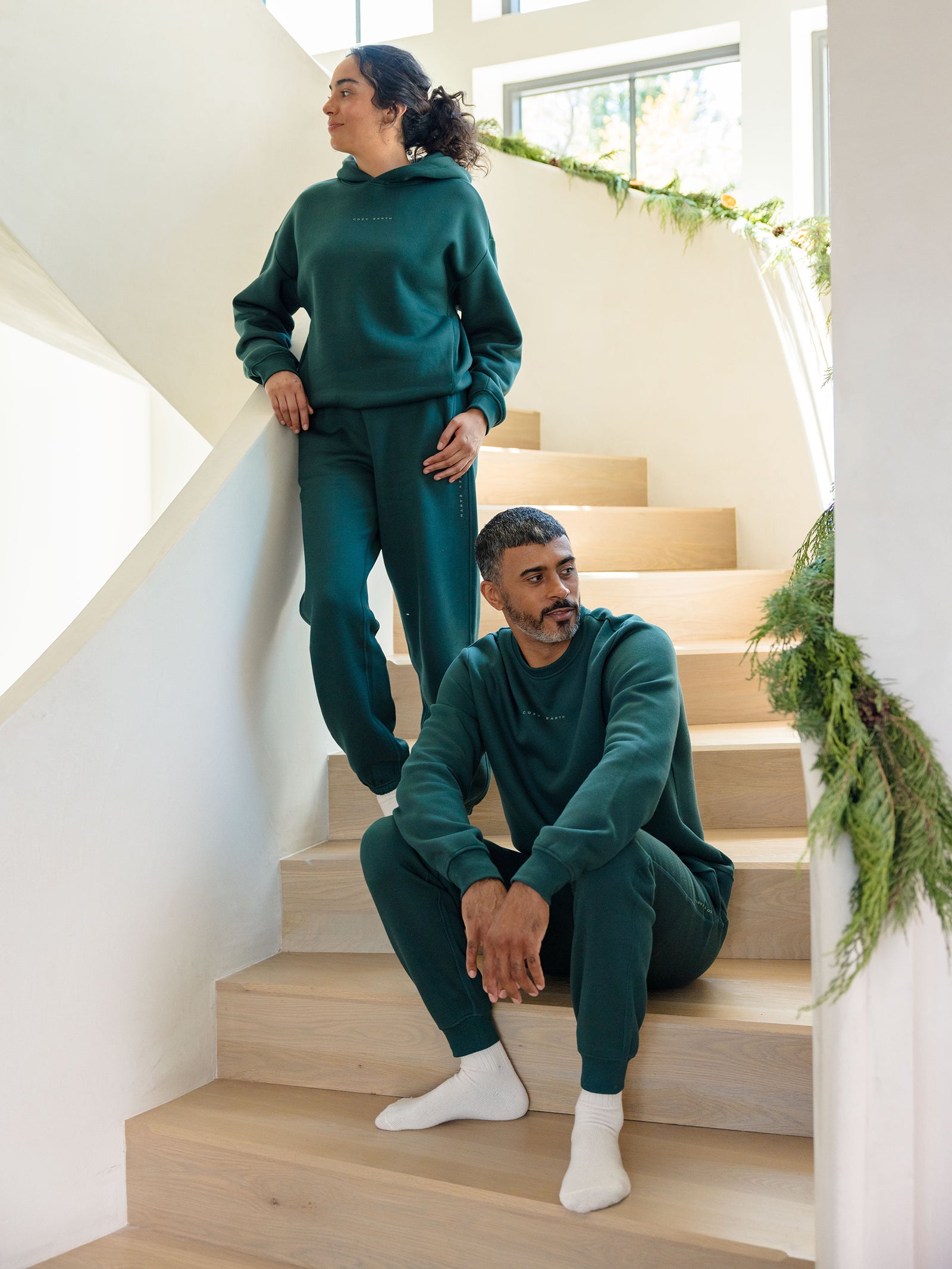 Two individuals, both donning the Men's CityScape Jogger by Cozy Earth in matching green, unwind on wooden stairs decorated with lush greenery. One person is seated on a step while the other stands with one foot on the step, gazing into the distance in a sunlit room. 