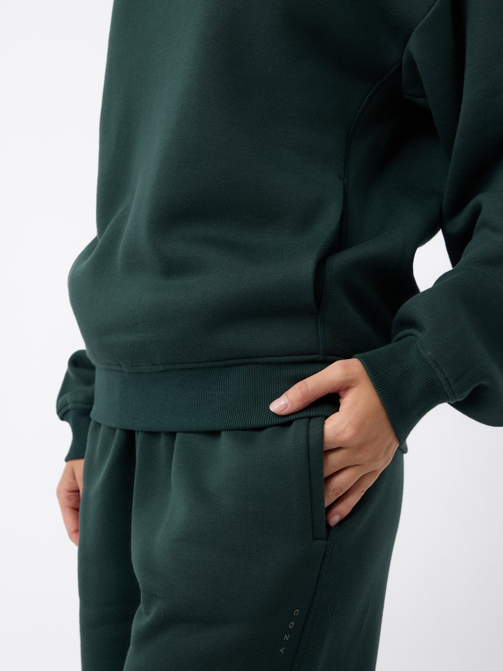 A person wearing a dark green tracksuit from Cozy Earth, featuring the Women's CityScape Sweatpant with their right hand in the pants pocket. The outfit includes a long-sleeved top and matching pants, both made from comfortable-looking fabric. The background is plain white. 