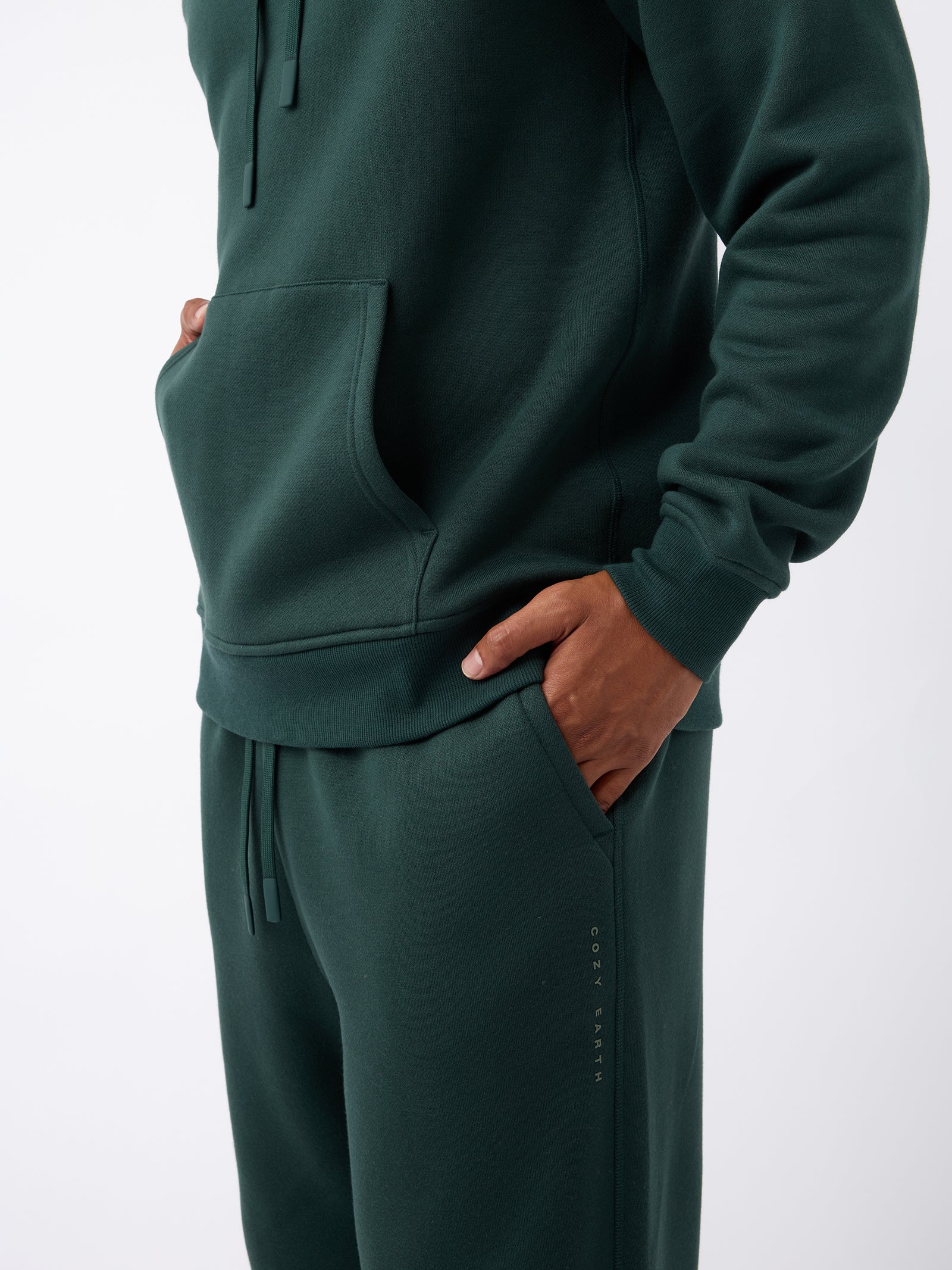 A person is wearing a dark green hoodie and matching Men's CityScape Sweatpants from Cozy Earth, with hands in pockets. The clothing features a subtle logo that reads "COZY EARTH" on the pants, set against a plain white background. 