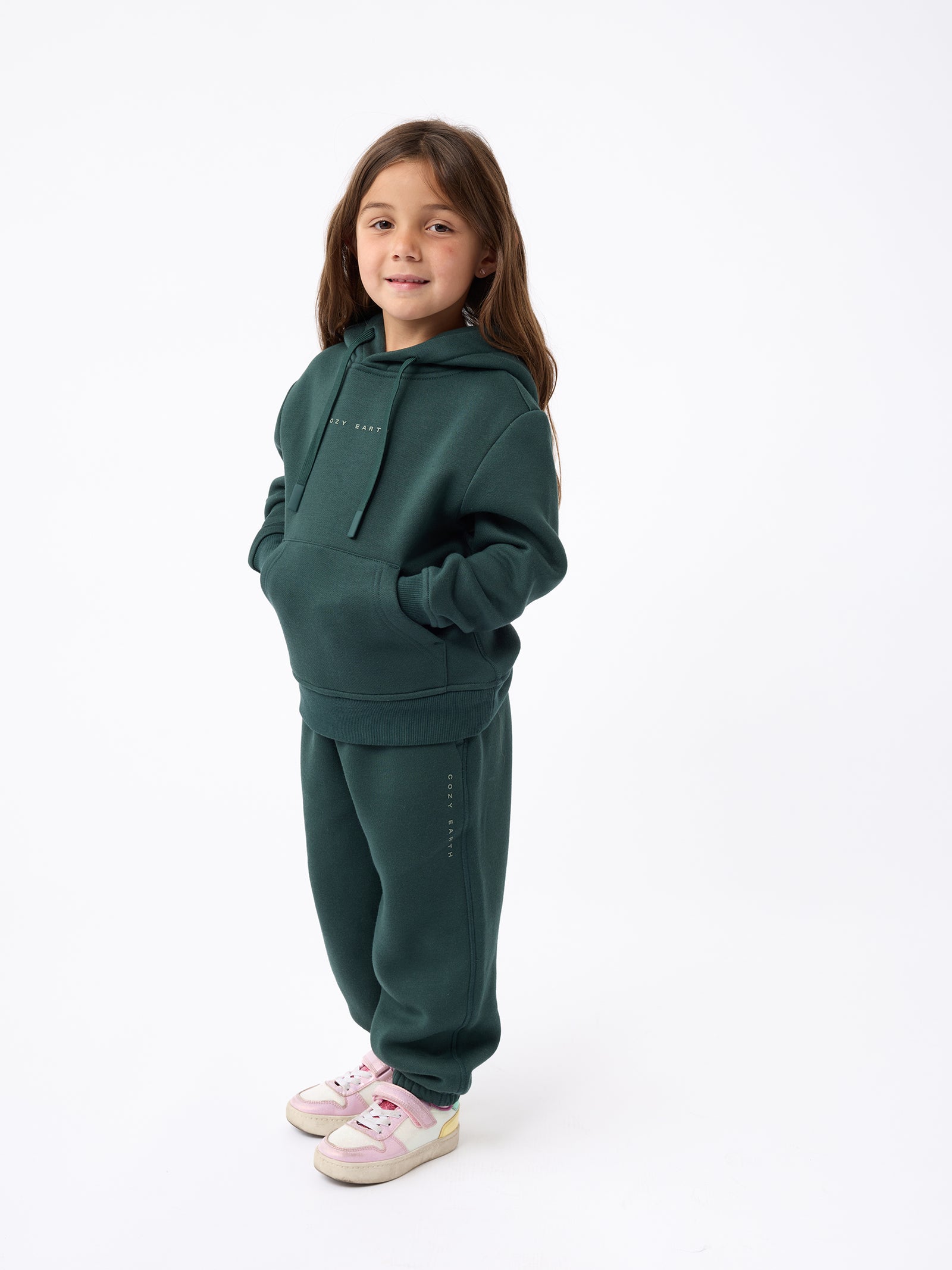 A young girl with long hair stands against a plain white background, wearing a dark green Cozy Earth hoodie and matching Kid's CityScape Sweatpant. Her hands are in her pockets, and she's sporting pink and white sneakers on her feet. 