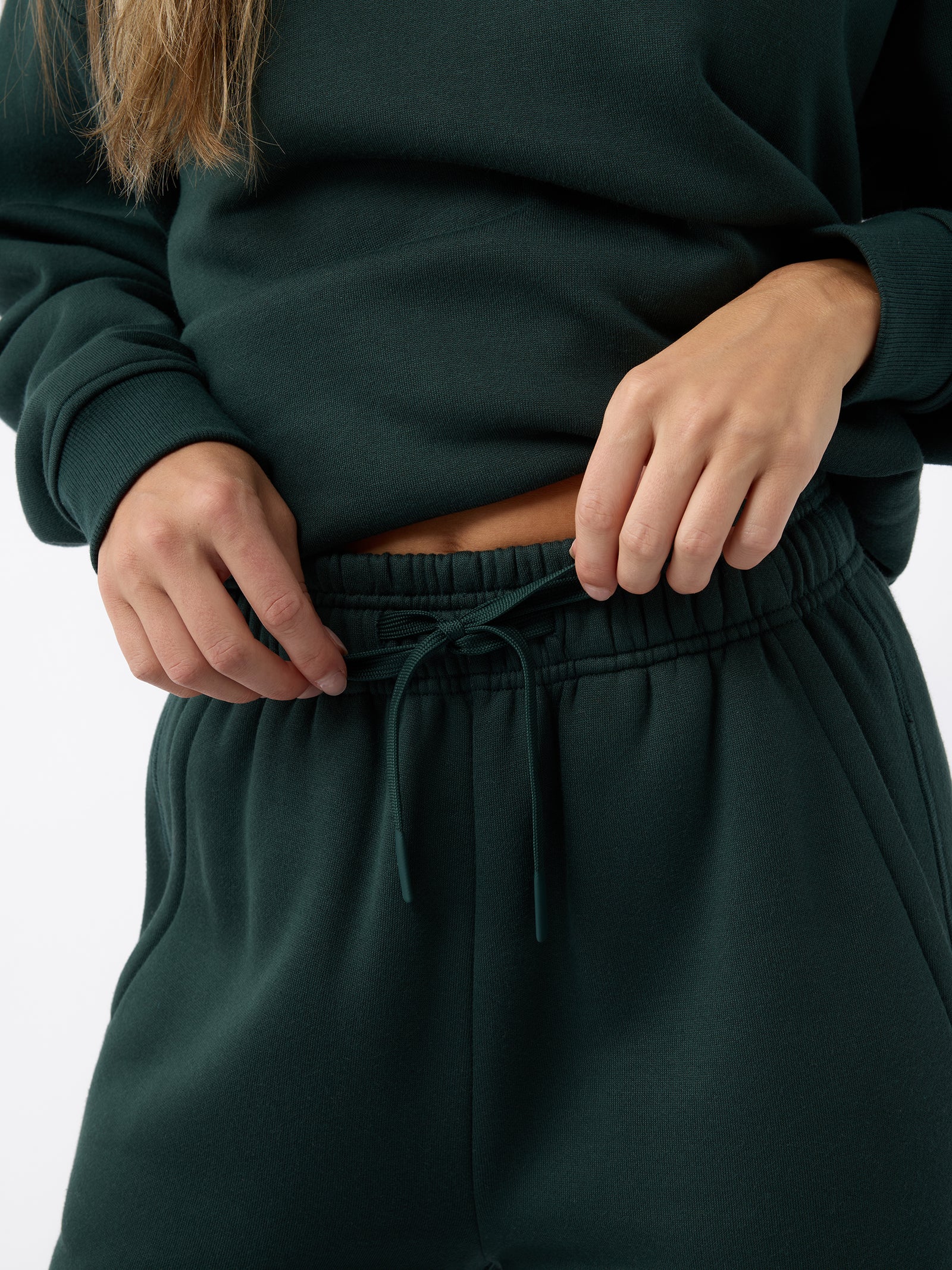 Close-up of a person adjusting the drawstring on Cozy Earth's Women's CityScape Wide Leg Pant in dark green. The individual is wearing a matching dark green sweatshirt against a plain white background. 