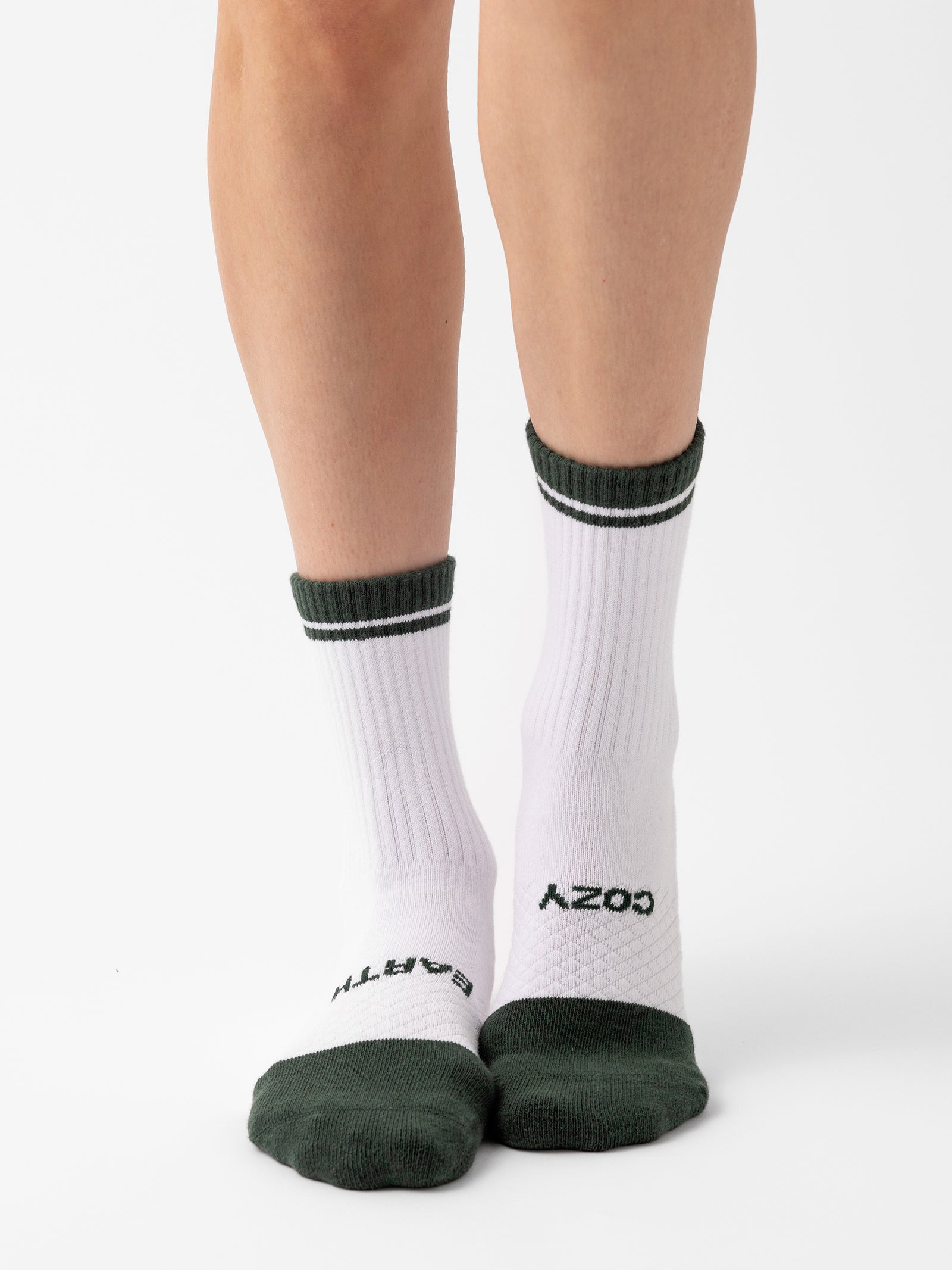 A pair of legs adorned with Essential Calf Socks from Cozy Earth, featuring white crew socks accented with dark green trim at the top, toes, and heels. The socks display "COZY" and "EARTH" in black text on the top of each foot. The background is plain white. 