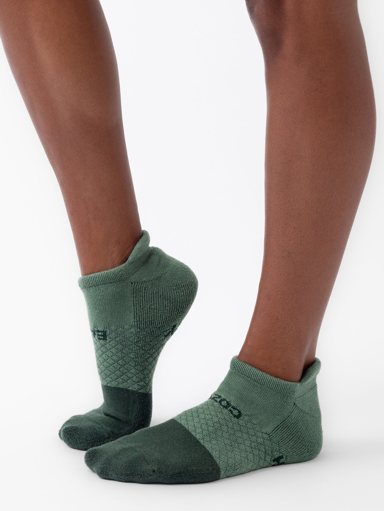 A person wearing green ankle-length socks from the Essential Ankle Sock 4-Pack by Cozy Earth, featuring a textured pattern with dark green accents on the toes and heels. The sides of the socks display the brand logo. The person is standing on tiptoe, accentuating the design and fit of these socks against a plain, white background. 