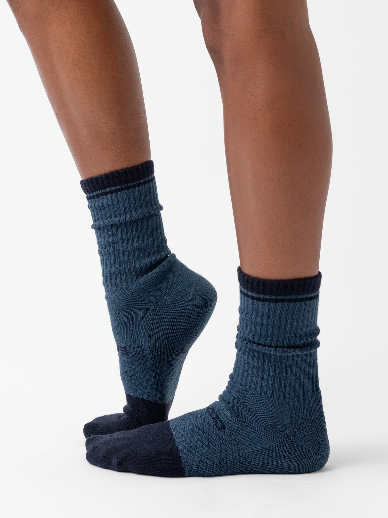 Close-up of a person's lower legs wearing a pair of dark blue Essential Calf Socks by Cozy Earth, featuring black toes, heels, and welts. The socks showcase ribbed cuffs and various textured knit patterns. The person is standing on the balls of their feet on a white background. 