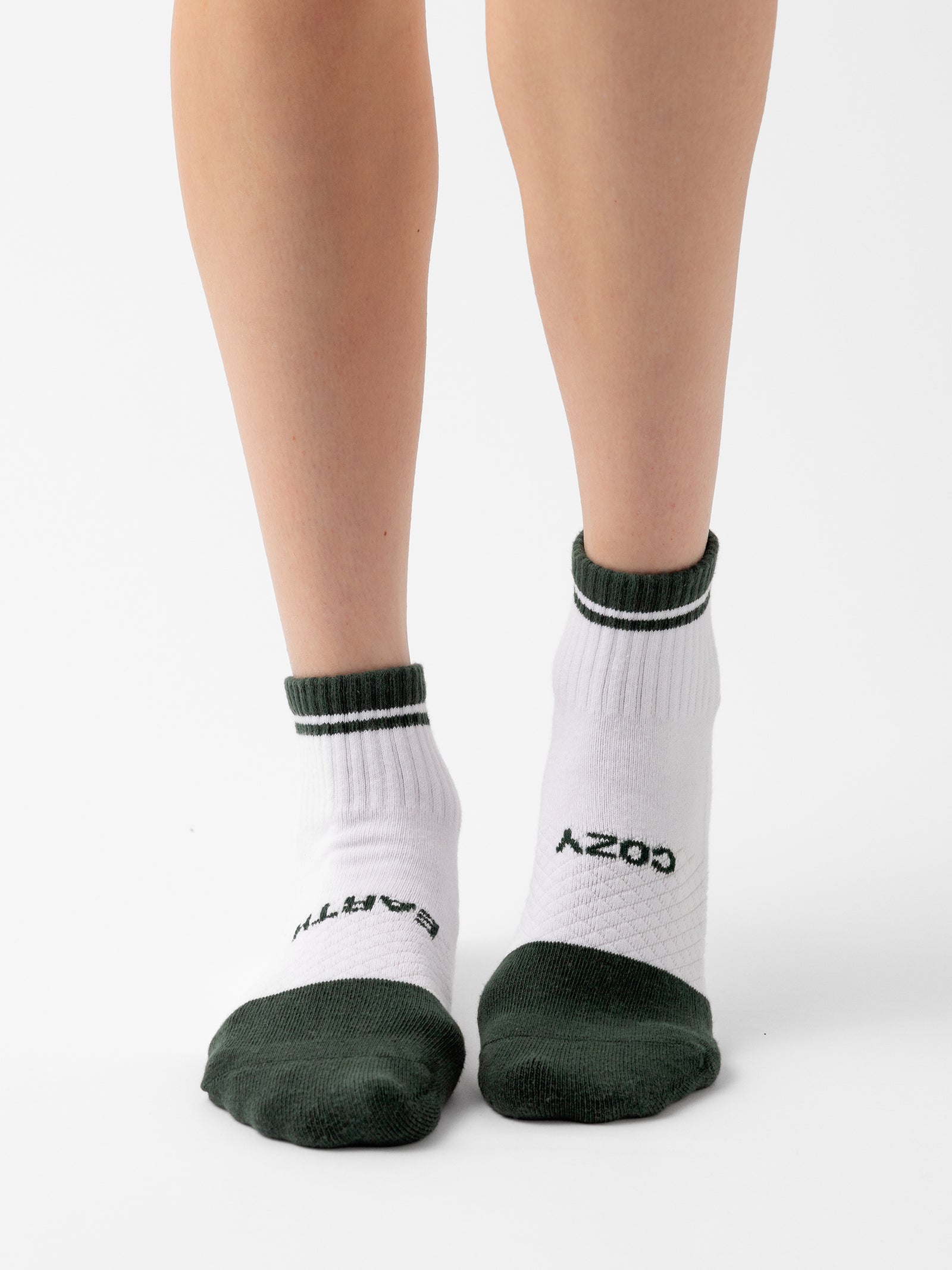 A pair of legs wearing white Essential Quarter Socks from Cozy Earth, featuring dark green accents on the toes, heels, and top trim. The socks have the words "COZY" and "CART" printed in black on the sides. The background is white. 