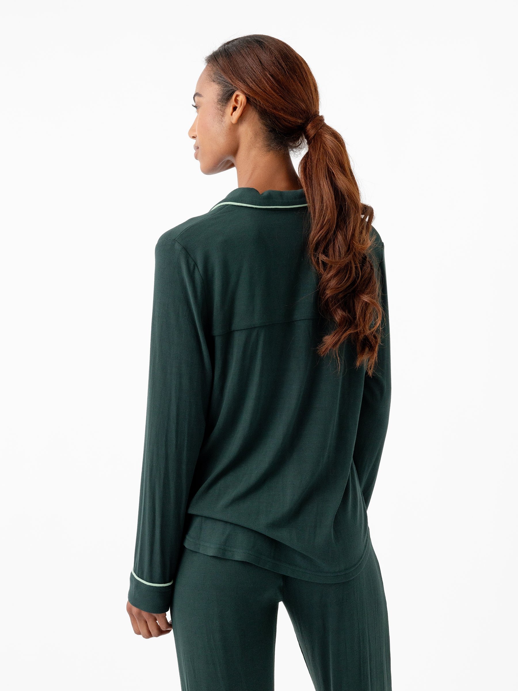 A person with long, wavy hair tied in a ponytail is standing and facing away from the camera. They are wearing a Cozy Earth Women's Bamboo Rib Knit Classic Long Sleeve Pajama Top with white trim on the cuffs and matching dark green pajama pants. The background is plain white. 