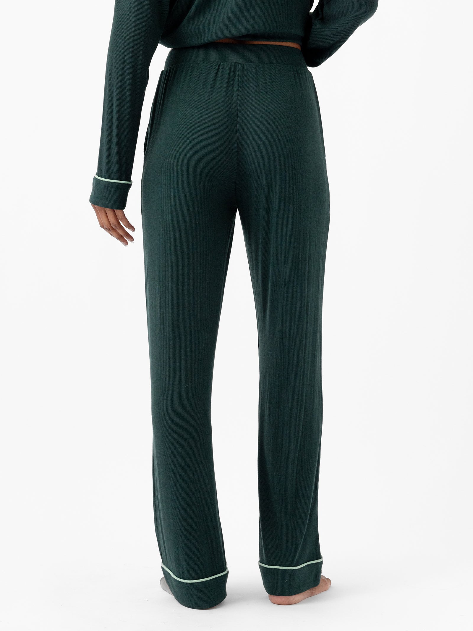 A person wearing Cozy Earth's dark green Women's Bamboo Rib Knit Classic Pajama Pant with white trim stands barefoot on a white background. The full-length pajama pants are complemented by the partially visible top of the matching long-sleeve shirt. The person is shown from the back. 