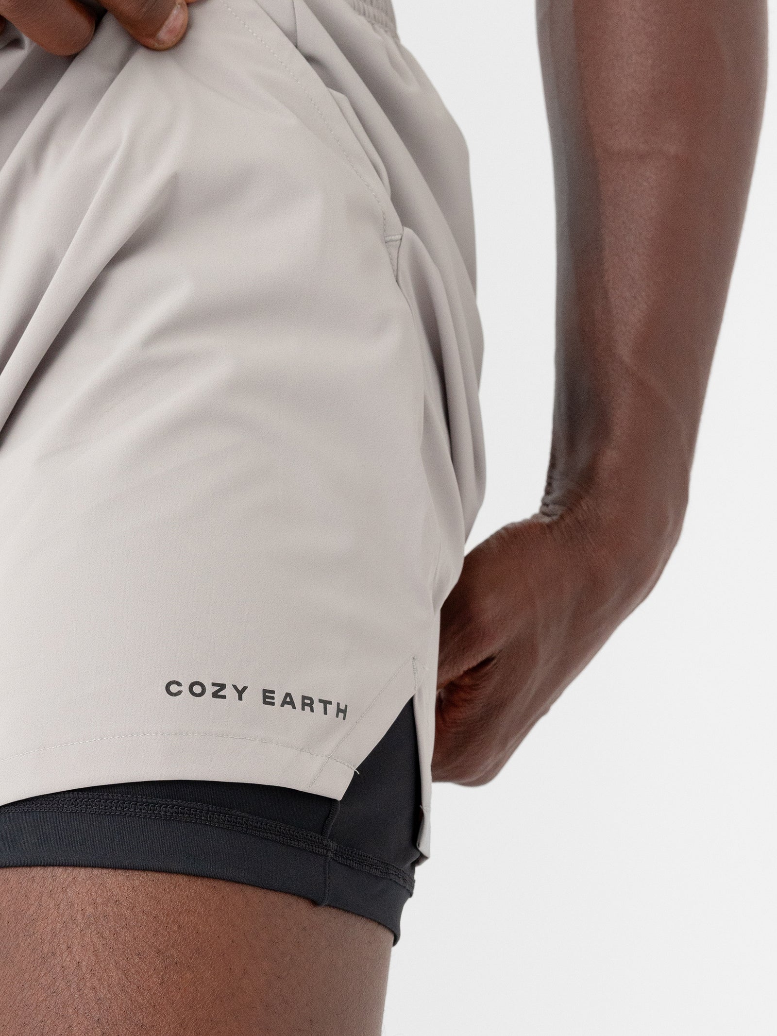 Close-up of a person's hand pulling light gray men's performance sleep shorts from Cozy Earth to the side, revealing black inner shorts. The person has a medium to dark skin tone, and the background is plain white. 