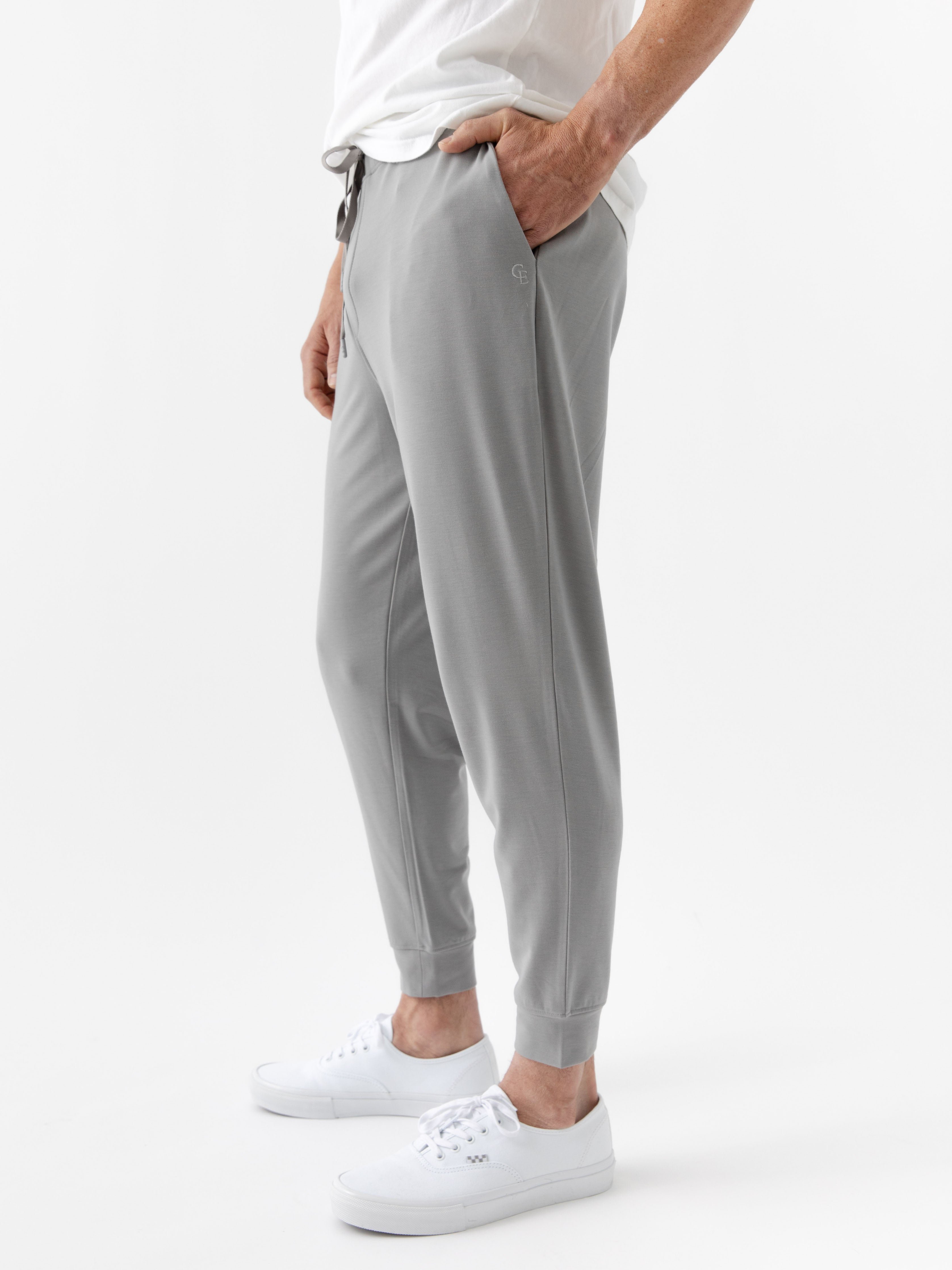 A person is wearing Cozy Earth's Men's Bamboo Jogger Set in gray with a white T-shirt, standing with one hand in their pocket and sporting white sneakers against a plain white background. 