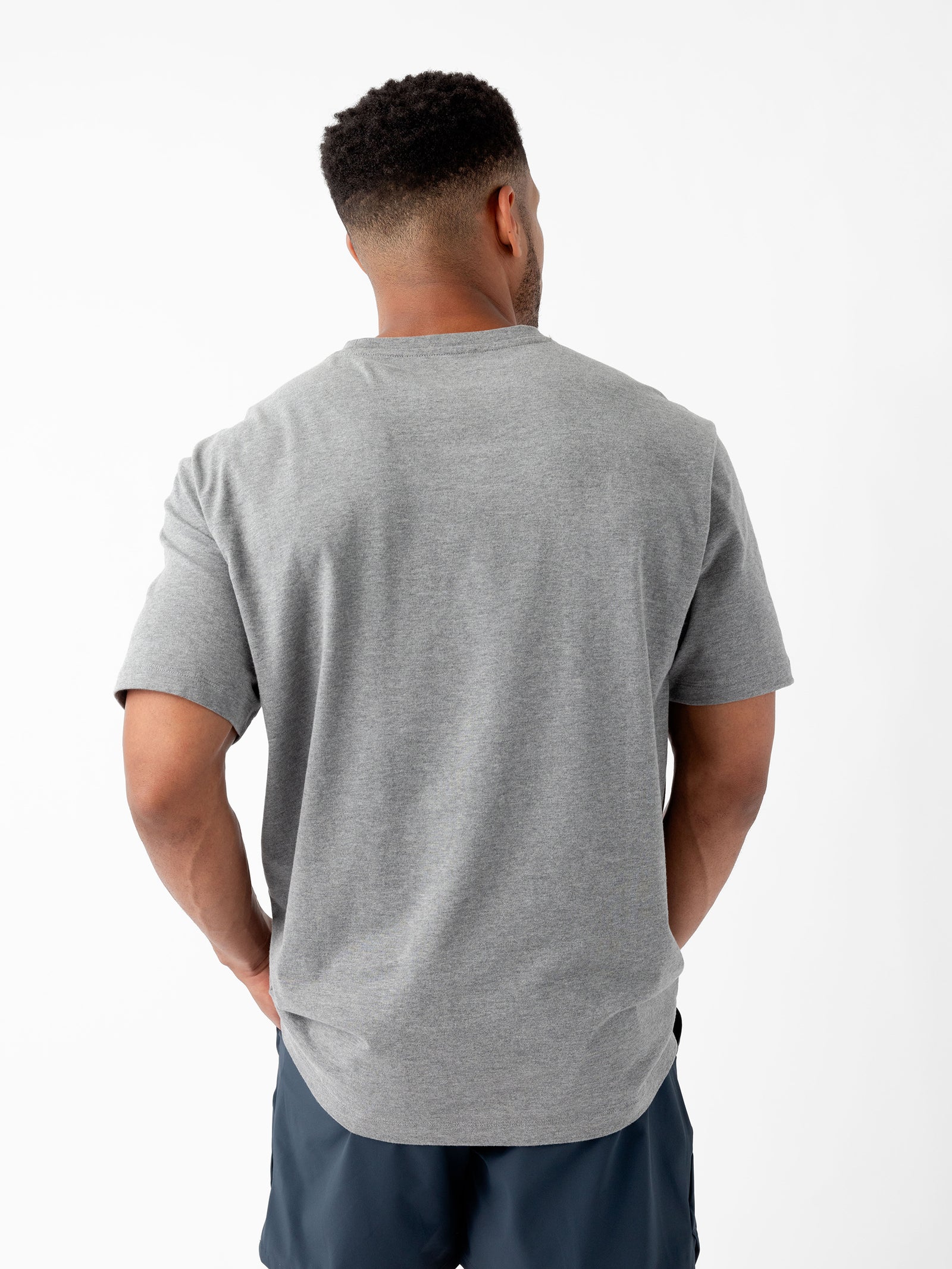 A person with short, dark curly hair is shown from the back, wearing a Cozy Earth Men's All Day Tee in gray and dark shorts. They stand against a plain white background. 