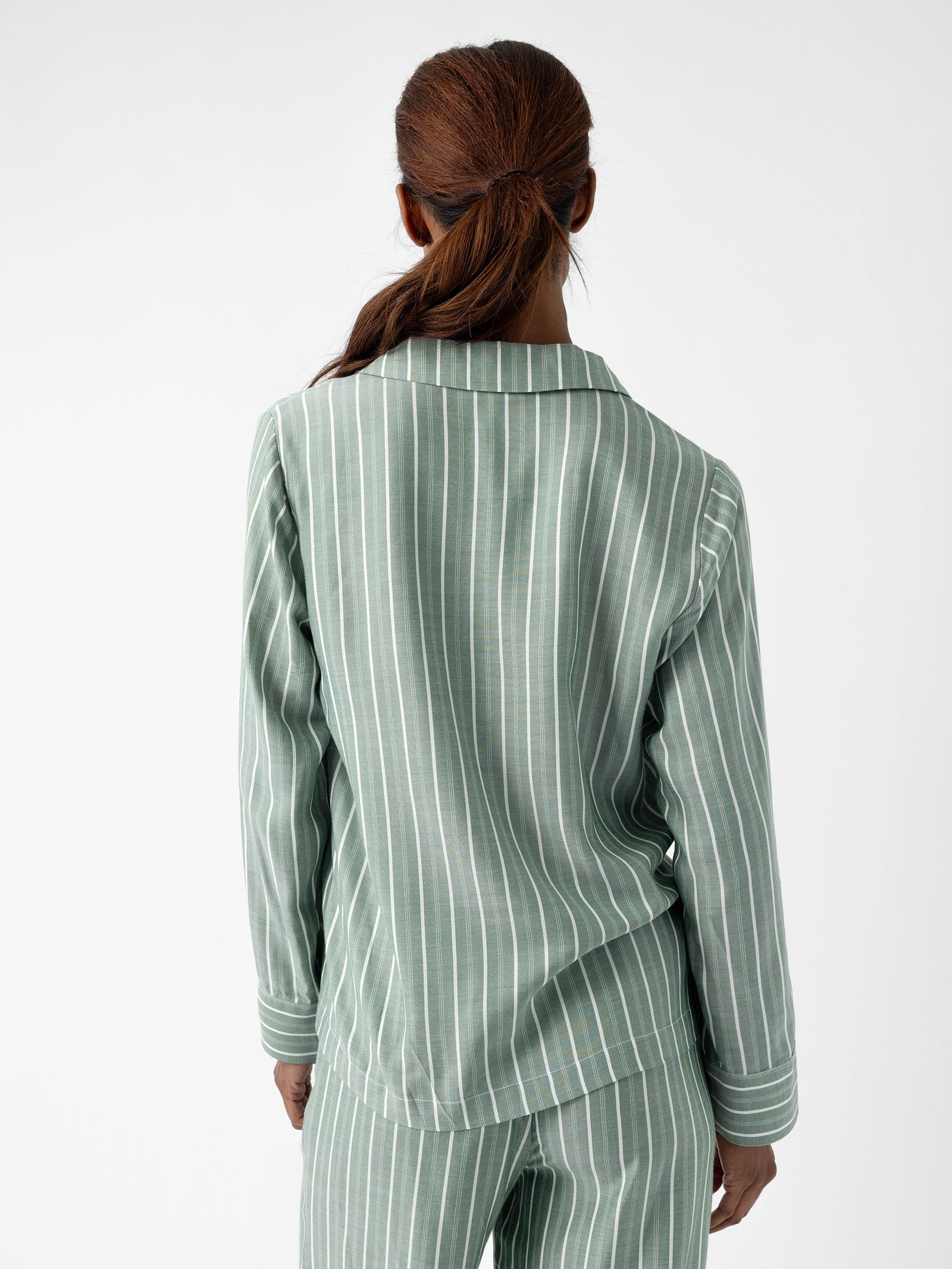 A woman with long brown hair is facing away from the camera, dressed in a Cozy Earth Women's Soft Woven Long Sleeve Pajama Top featuring matching green and white vertical stripes. She stands against a plain white background. 