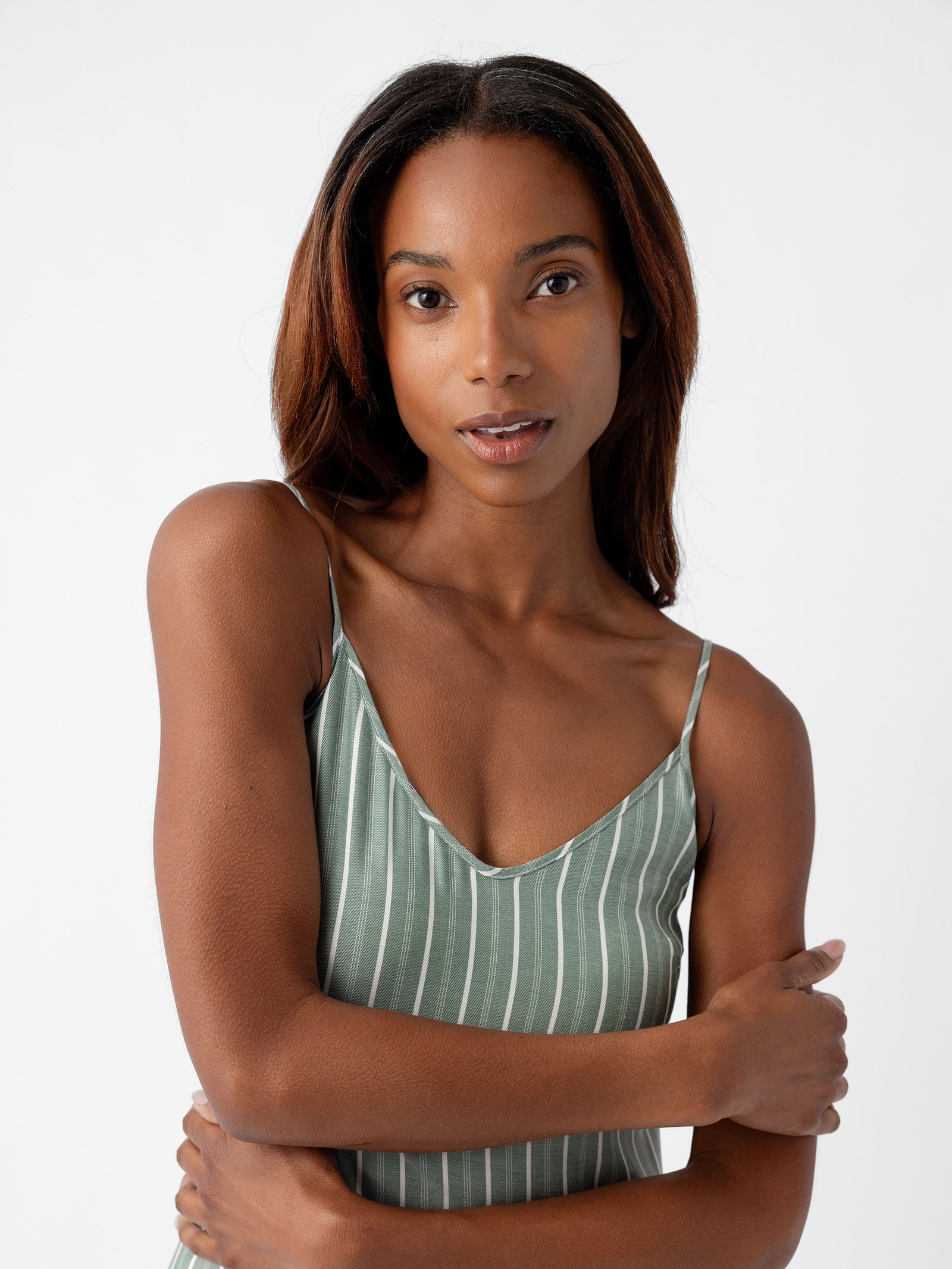 A woman with long hair, dressed in a green and white striped spaghetti strap top from Cozy Earth's Women's Soft Woven Nightgown collection, stands against a plain background, crossing her arms and looking at the camera. 