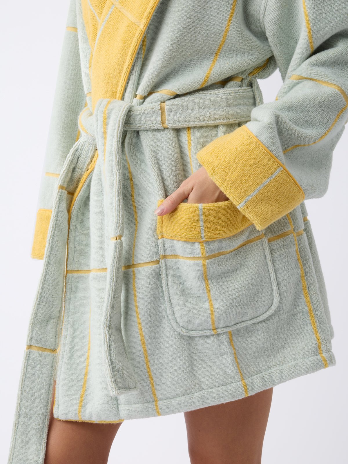 A person in a Cozy Earth Windowpane Resort Robe, showcasing a yellow and green checkered pattern with large pockets and a matching belt, stands with one hand in a pocket against a plain white background. 