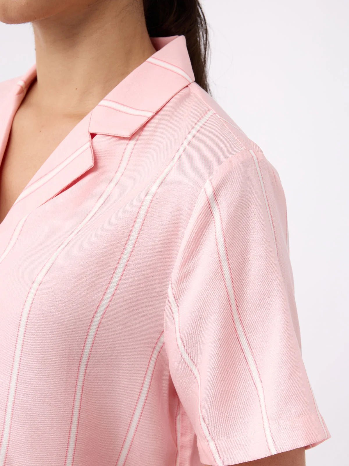 A close-up shows a person wearing the Women's Soft Woven Short Sleeve Pajama Top by Cozy Earth, featuring a pink color with white stripes, collar, and short sleeves against a plain white background. 