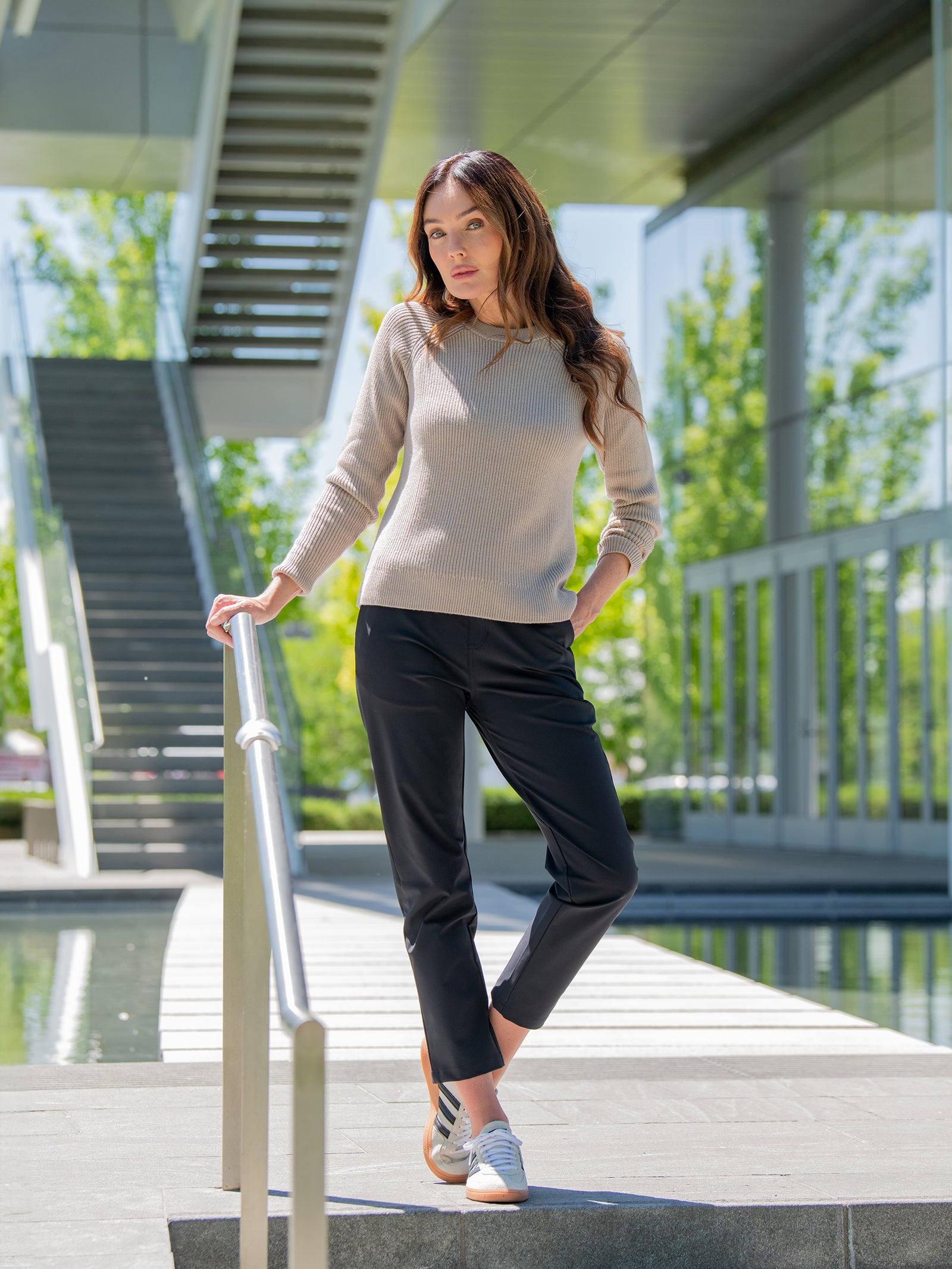 A person in Cozy Earth's Women's Classic Crewneck and black pants stands outdoors on a walkway by a railing. They have one hand on the railing and the other in their pocket. They are under a modern building structure featuring glass and metal elements, with greenery visible around. 