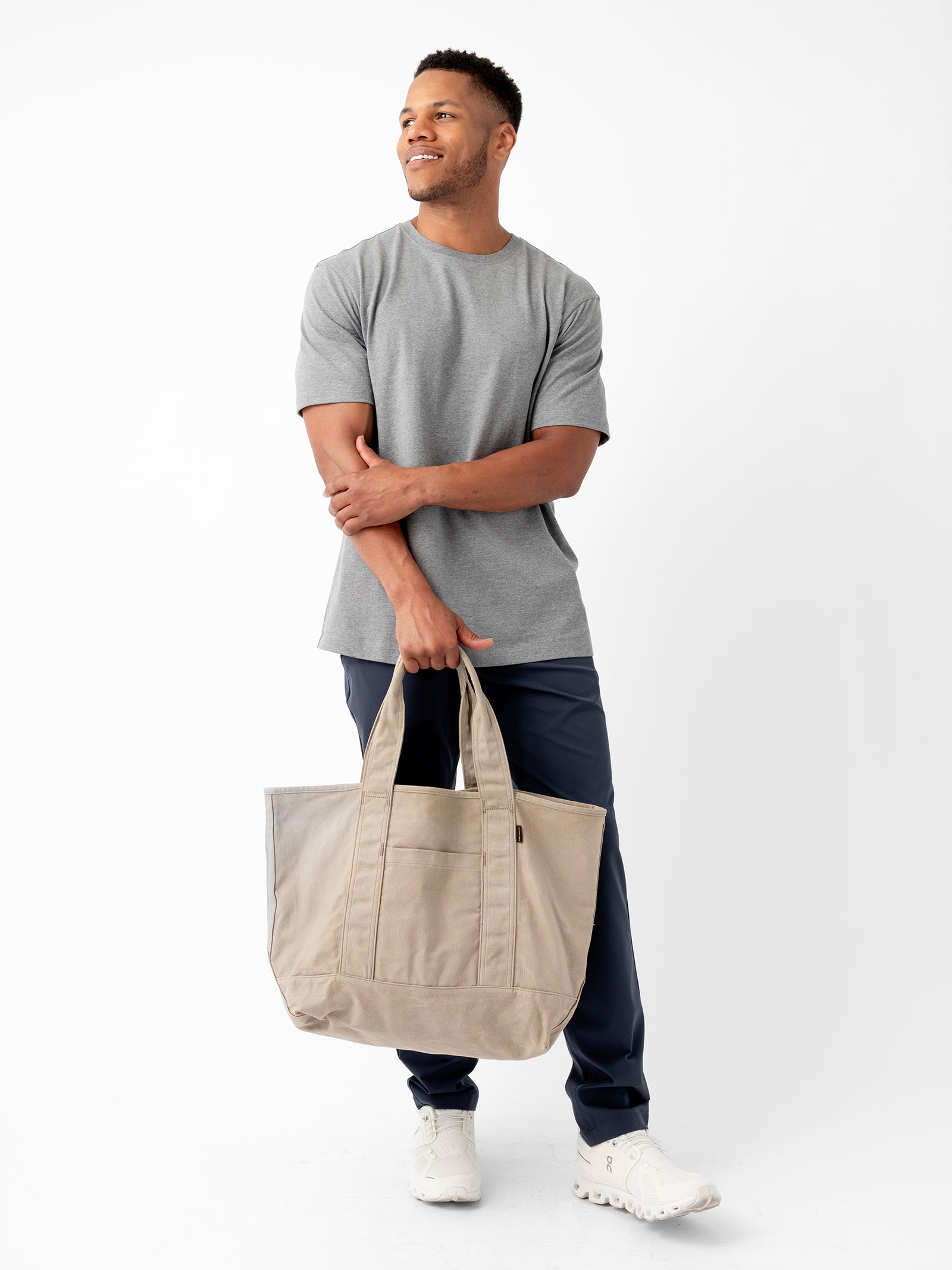 A man stands against a plain white background, smiling slightly while looking off to the side. He is wearing a gray T-shirt, dark blue pants, and white sneakers, and is holding the Waxed Canvas Tote from Cozy Earth. His arms are crossed casually as he carries the bag. 