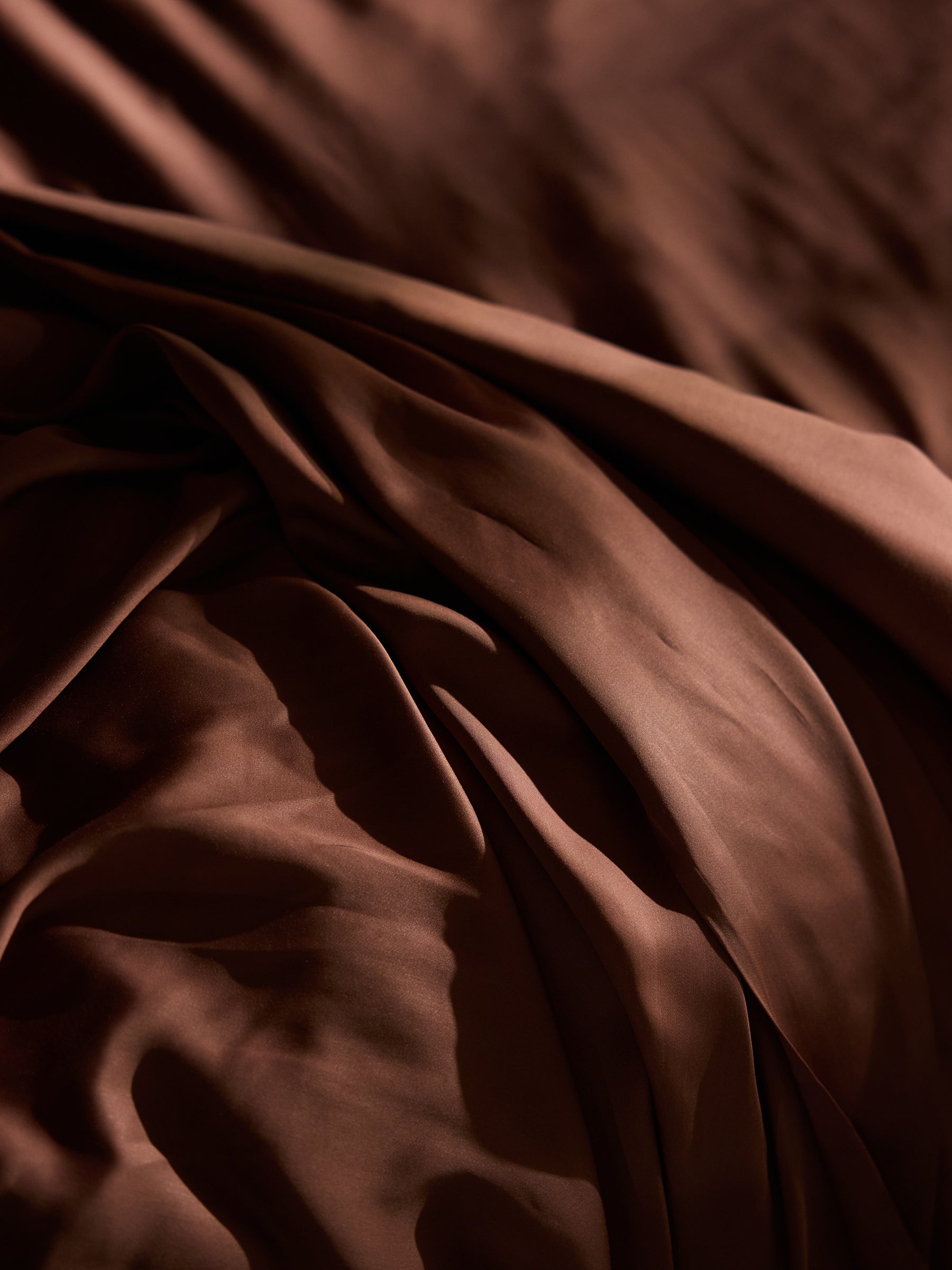 A close-up shot of smooth, rumpled brown satin fabric in soft lighting. The material has gentle waves and folds, creating a luxurious and elegant texture. Shadows and highlights add depth to the Bamboo Sheet Set by Cozy Earth's rich surface. 