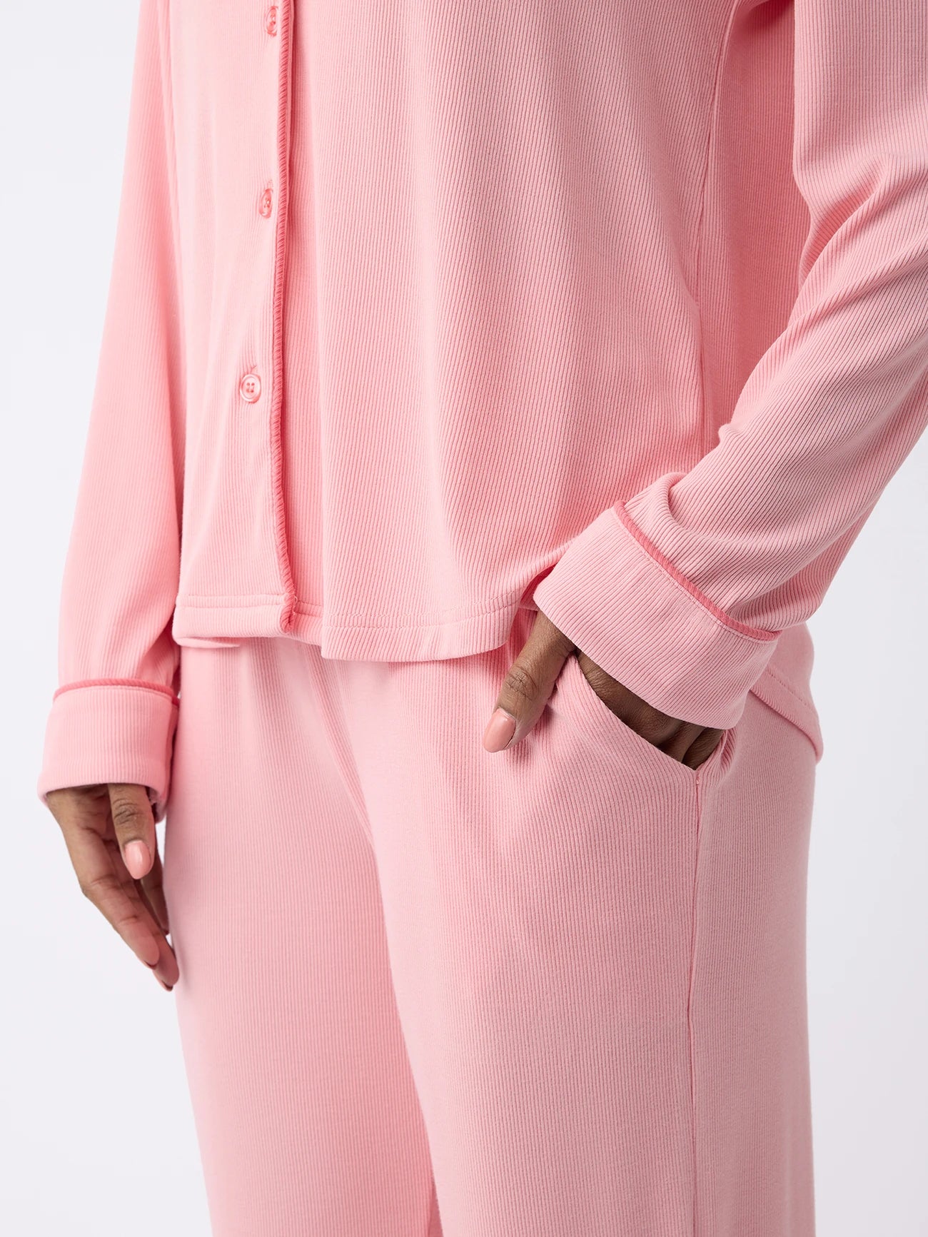 A woman is shown wearing Cozy Earth's Women's Bamboo Rib-Knit Classic Pajama Pant in soft pink, featuring a ribbed texture and long sleeves. Their hand rests in the pants pocket against a plain white background. 