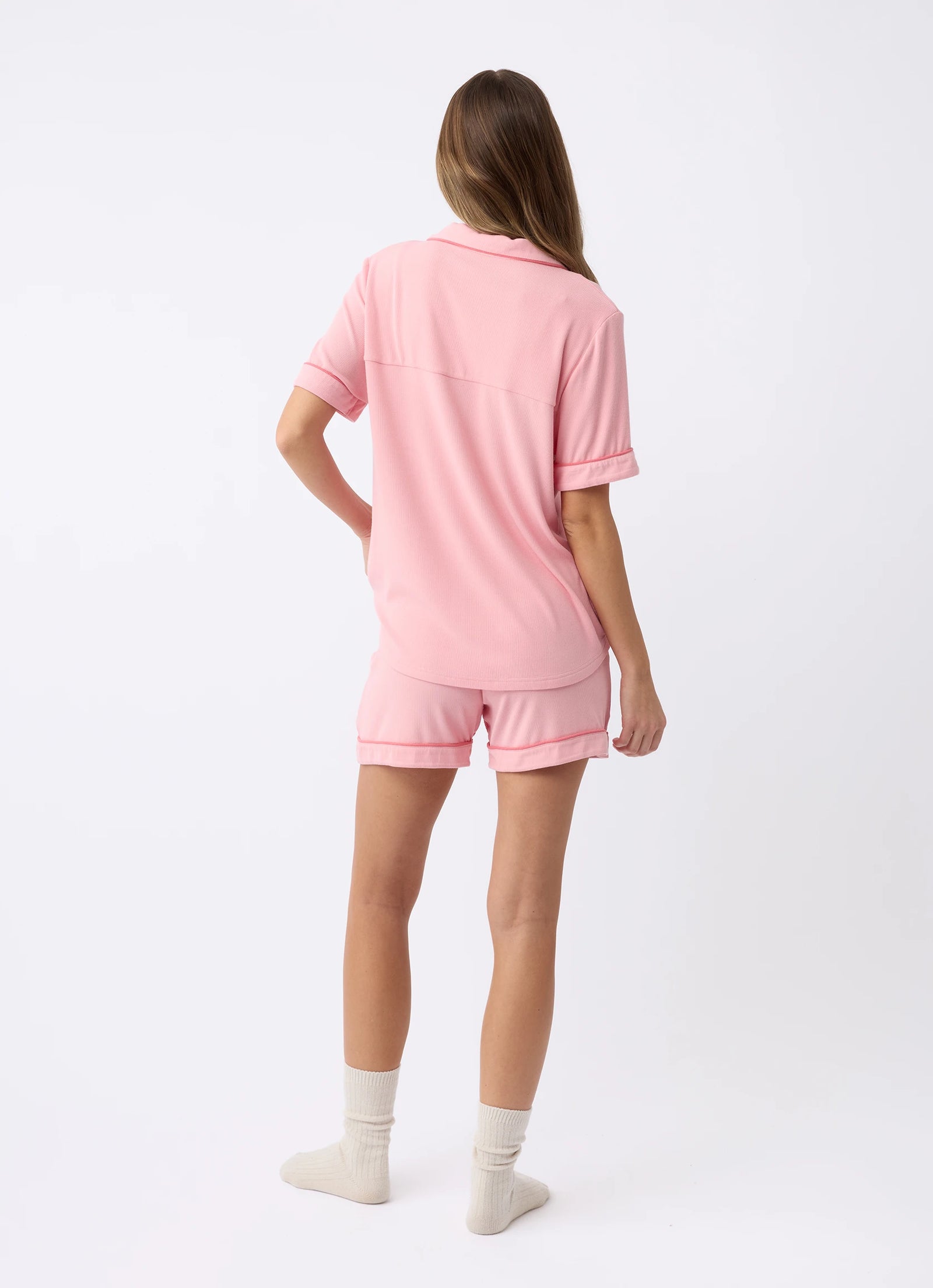 A woman with long hair is facing away, barefoot, wearing a casual pink shirt and Women's Bamboo Rib-Knit Classic Pajama Short by Cozy Earth. The background is plain white. 