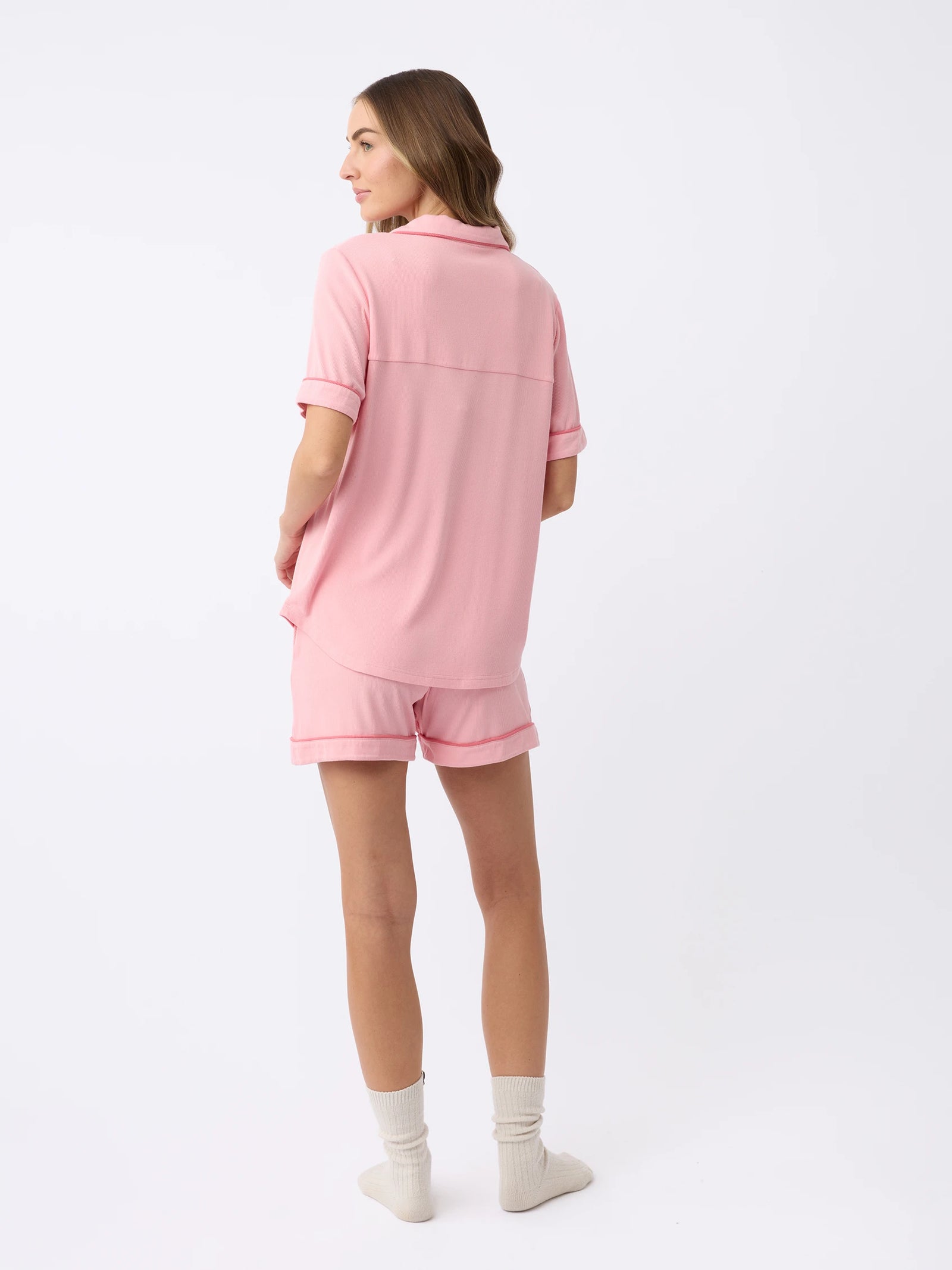 A woman stands with her back turned, wearing Cozy Earth's Women's Bamboo Rib-Knit Classic Short Sleeve Pajama Top and matching shorts. She is standing on a white background, wearing white socks and looking to the side. 