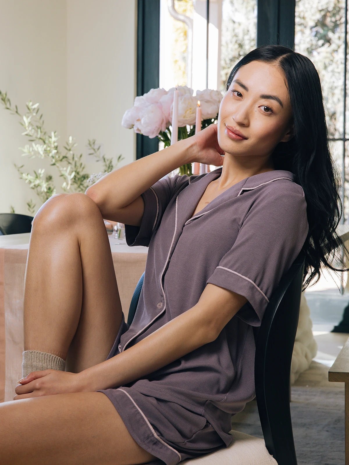 A woman with long dark hair sits comfortably in a sunlit room, wearing the Cozy Earth Women's Rib-Knit Classic Short Sleeve Pajama Set. One hand rests on her neck while pink flowers and greenery decorate the area near a large window. 